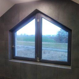 WW-REM - Okno dwuskrzydłowe w kształcie trapezu, osadzone w ścianie wyłożonej ciemnymi, kwadratowymi płytkami. Widok na zewnątrz ukazuje zielone pola i drzewa.