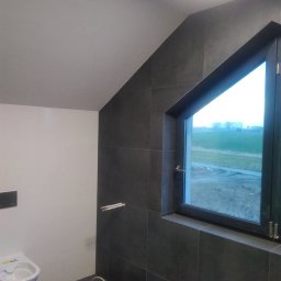 WW-REM - Ciemne płytki w łazience na poddaszu z oknem, widoczna toaleta i akcesoria montażowe. Ukończone wykończenie wnętrza z nietypowym oknem.