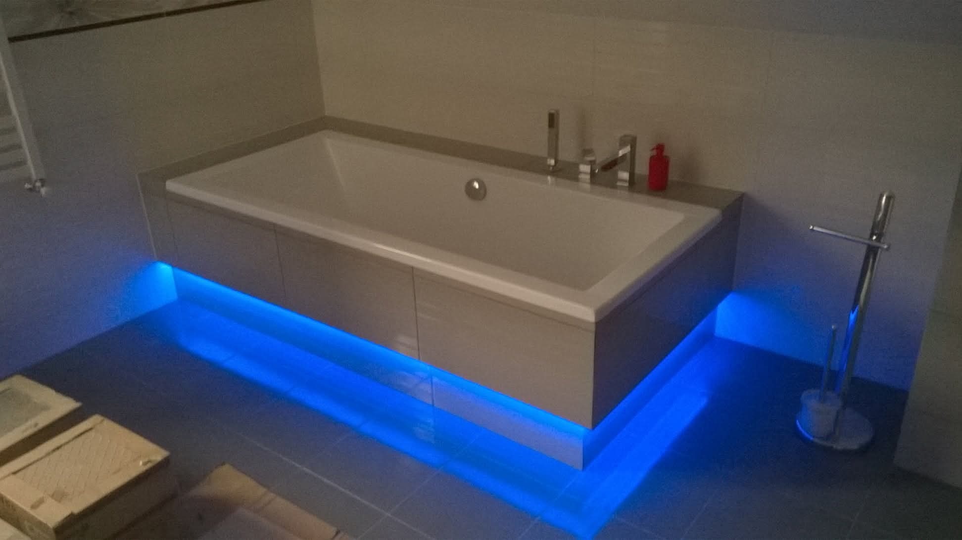 Nowoczesna wanna z podświetleniem LED w odcieniach błękitu, umieszczona w minimalistycznej łazience z szarymi płytkami. Widoczny chromowany zestaw baterii i akcesoria.