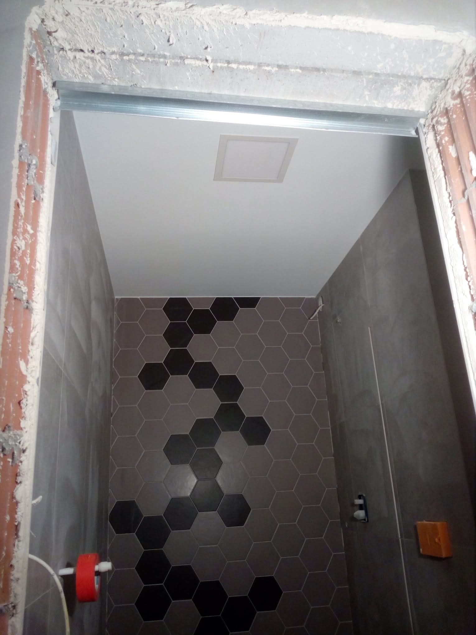 Wnętrze łazienki w trakcie wykańczania: szare płytki heksagonalne z czarnymi akcentami na ścianie, sufit podwieszany z oświetleniem, widoczne prace wykończeniowe przy futrynie.