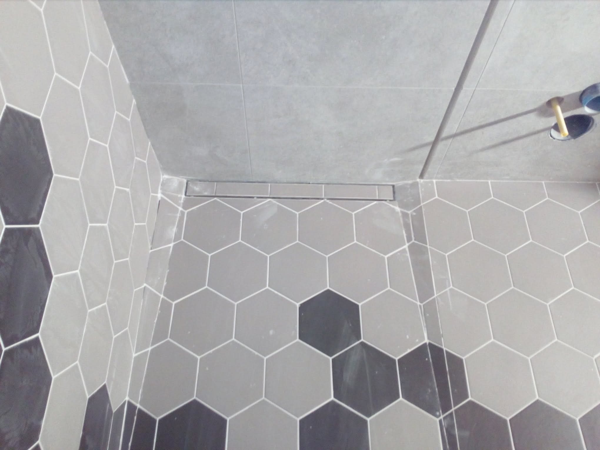 Wykończona łazienka z heksagonalnymi płytkami w odcieniach szarości i czerni, widoczny odpływ liniowy i fragment instalacji. Minimalistyczny design.