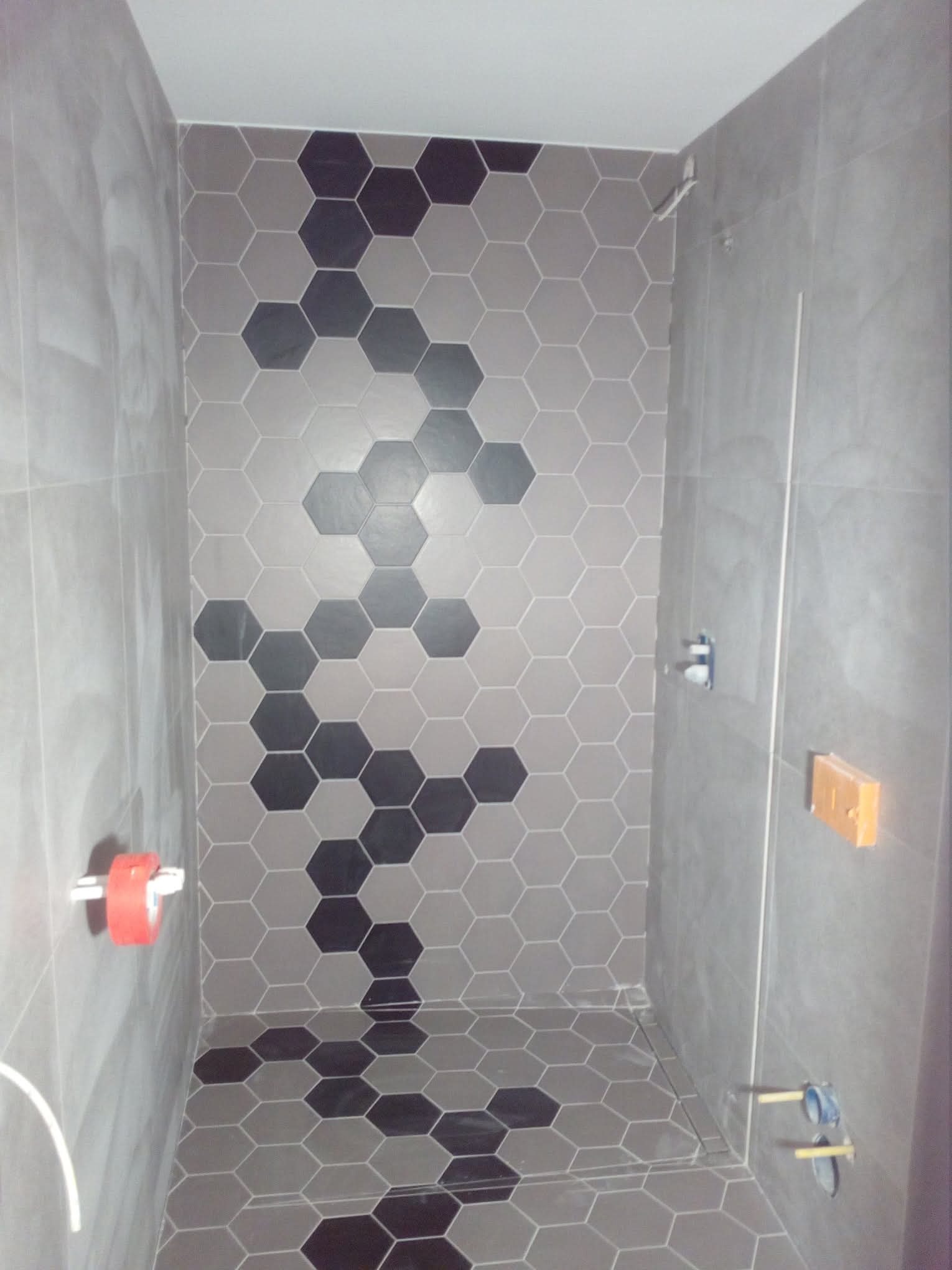 Wykończona łazienka z szarymi, heksagonalnymi płytkami, tworzącymi wzór. Widoczne elementy instalacji wodnej i akcesoria łazienkowe. Minimalistyczny design.