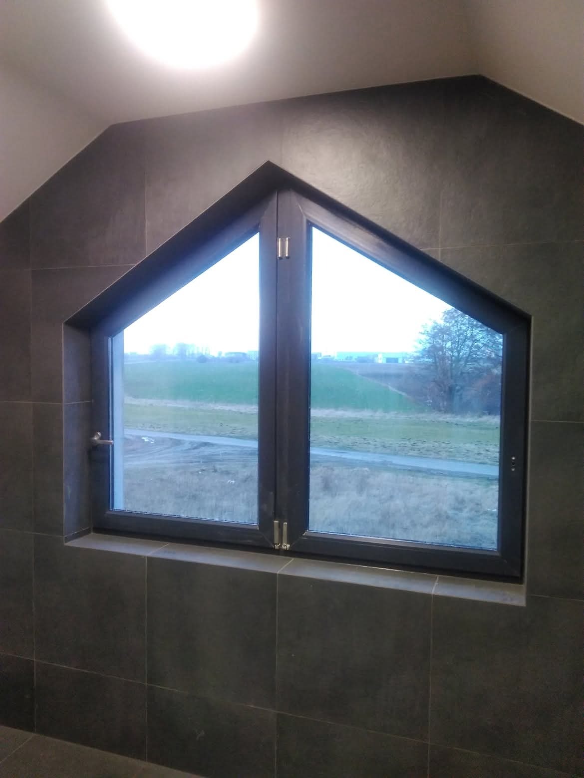 Okno dwuskrzydłowe w kształcie trapezu, osadzone w ścianie wyłożonej ciemnymi, kwadratowymi płytkami. Widok na zewnątrz ukazuje zielone pola i drzewa.