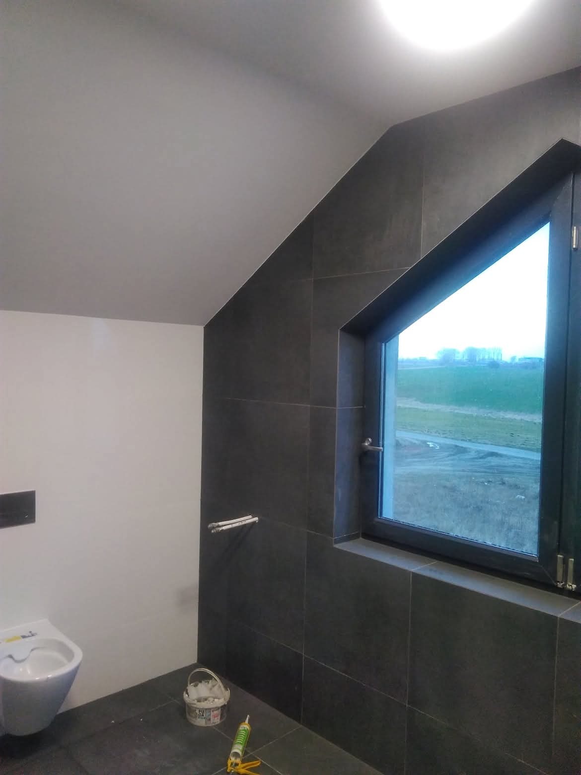 Ciemne płytki w łazience na poddaszu z oknem, widoczna toaleta i akcesoria montażowe. Ukończone wykończenie wnętrza z nietypowym oknem.