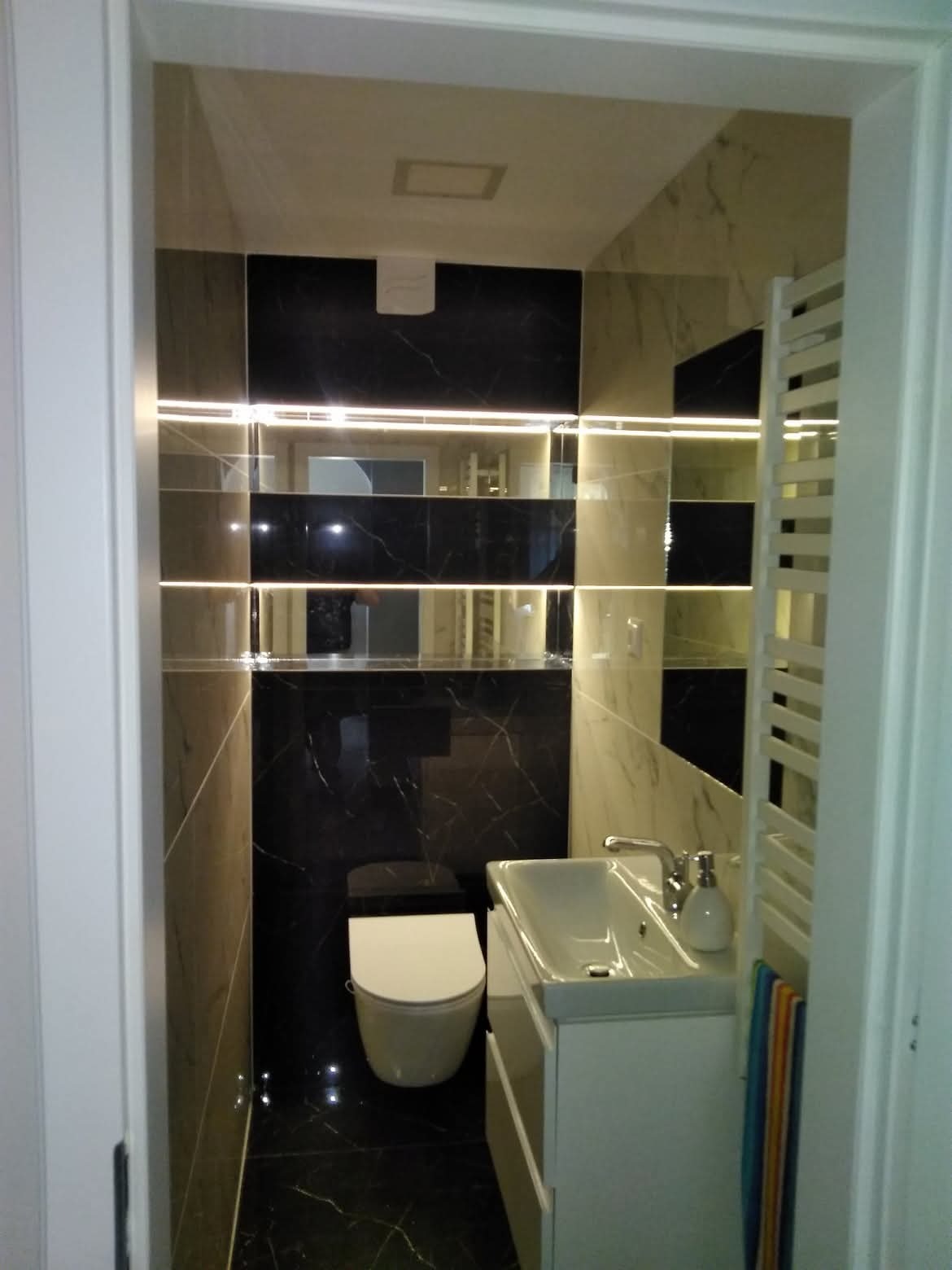 Nowoczesna łazienka z czarno-białymi marmurowymi płytkami, podświetlanymi półkami i minimalistyczną armaturą. Widok zza futryny drzwi.