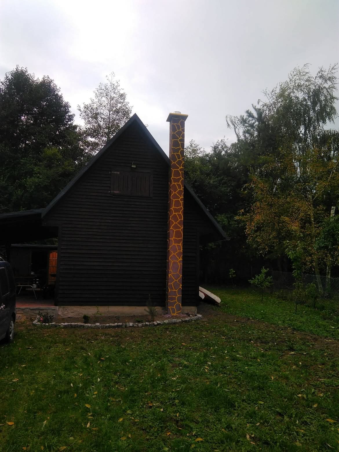 Ciemna, drewniana chatka z wysokim kominem w brązowo-żółte wzory, otoczona zielenią. Komin wyróżnia się na tle ciemnego drewna i dodaje unikalnego charakteru budynkowi.