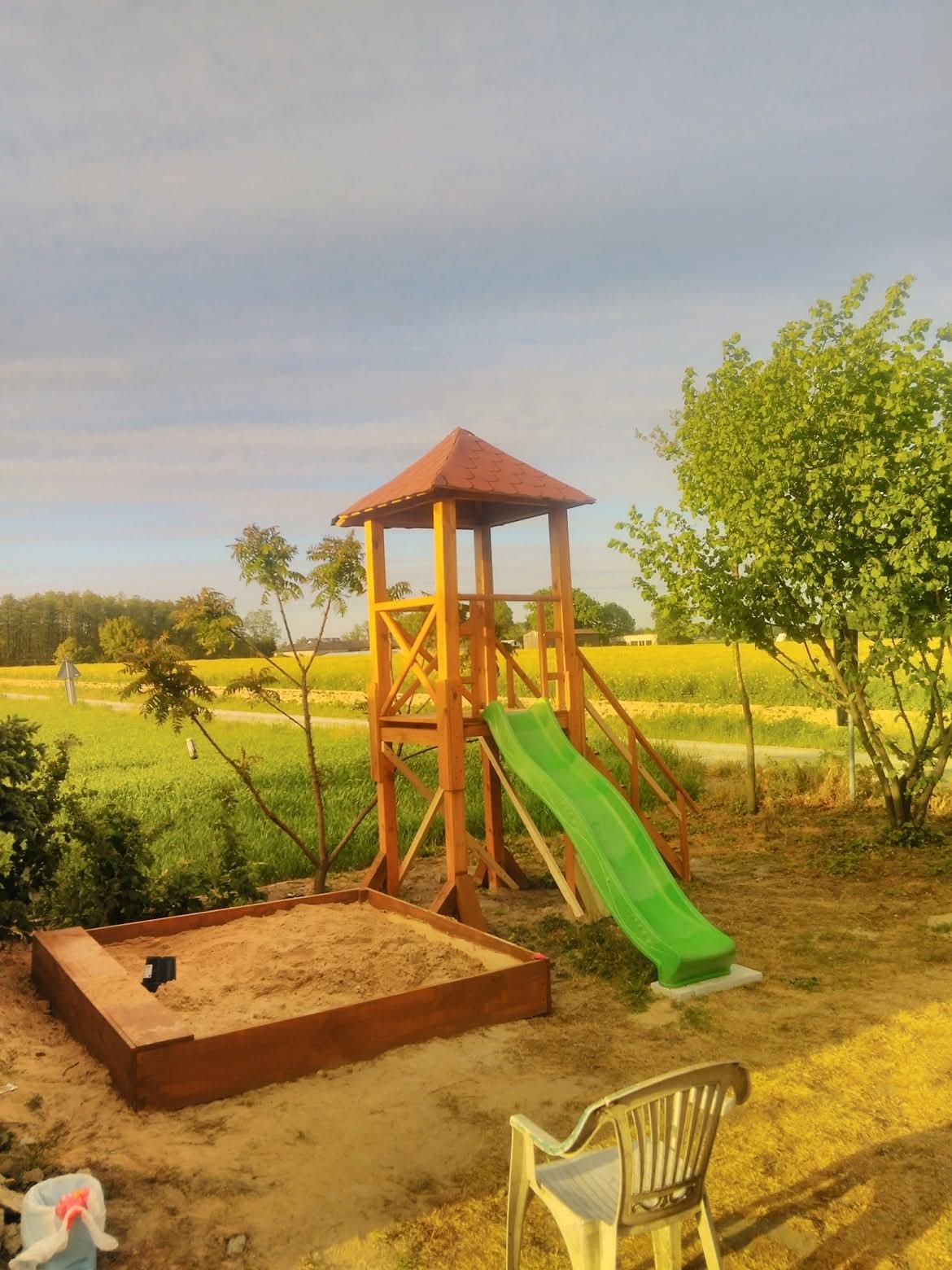 Drewniany plac zabaw z wieżą, zjeżdżalnią i piaskownicą na tle zielonego pola i błękitnego nieba. Widoczny plastikowy krzesło.