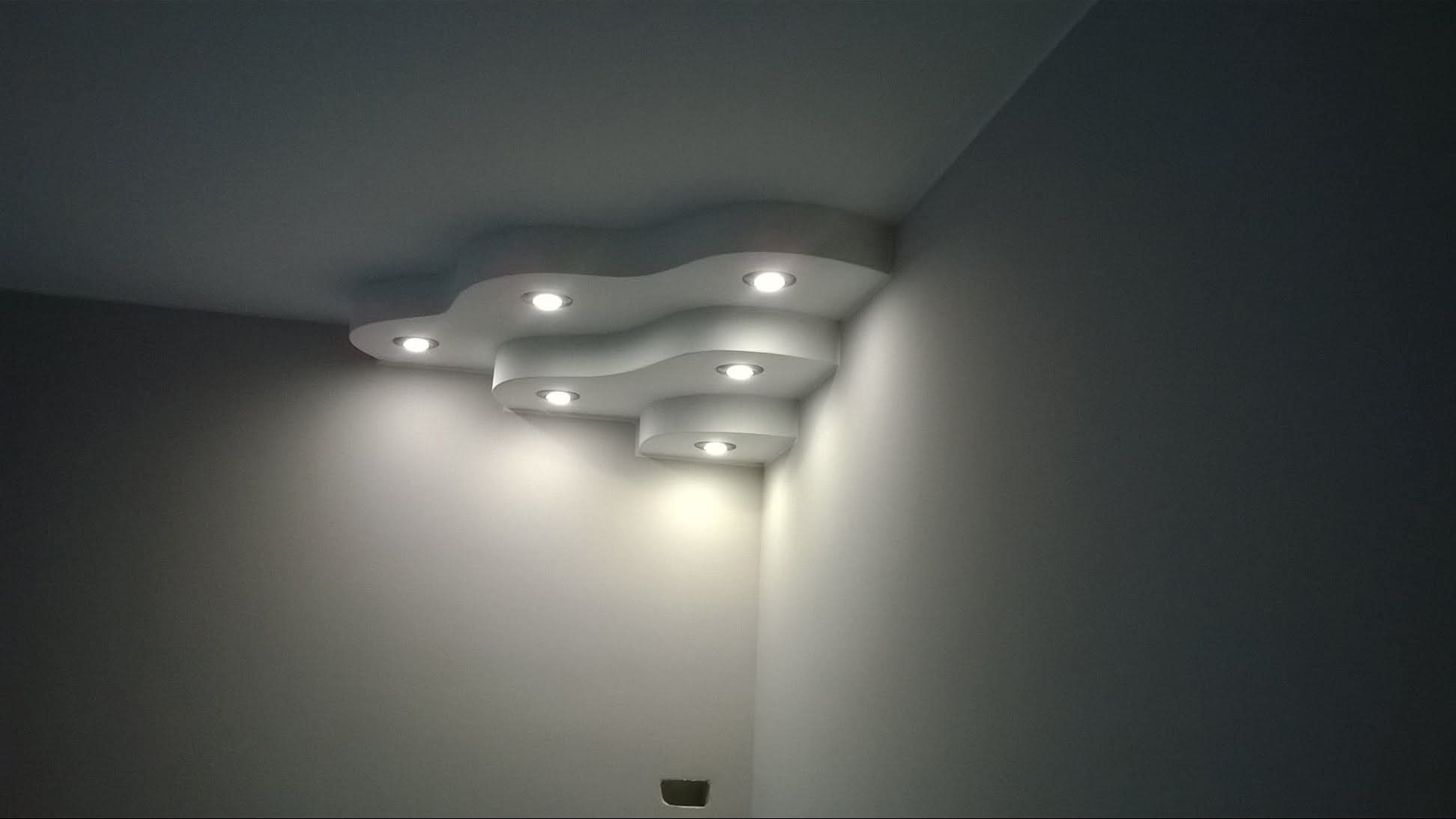 Nowoczesny sufit podwieszany z oświetleniem punktowym w rogu pokoju. Białe ściany, minimalistyczny design. Widoczne delikatne cienie i gra świateł.