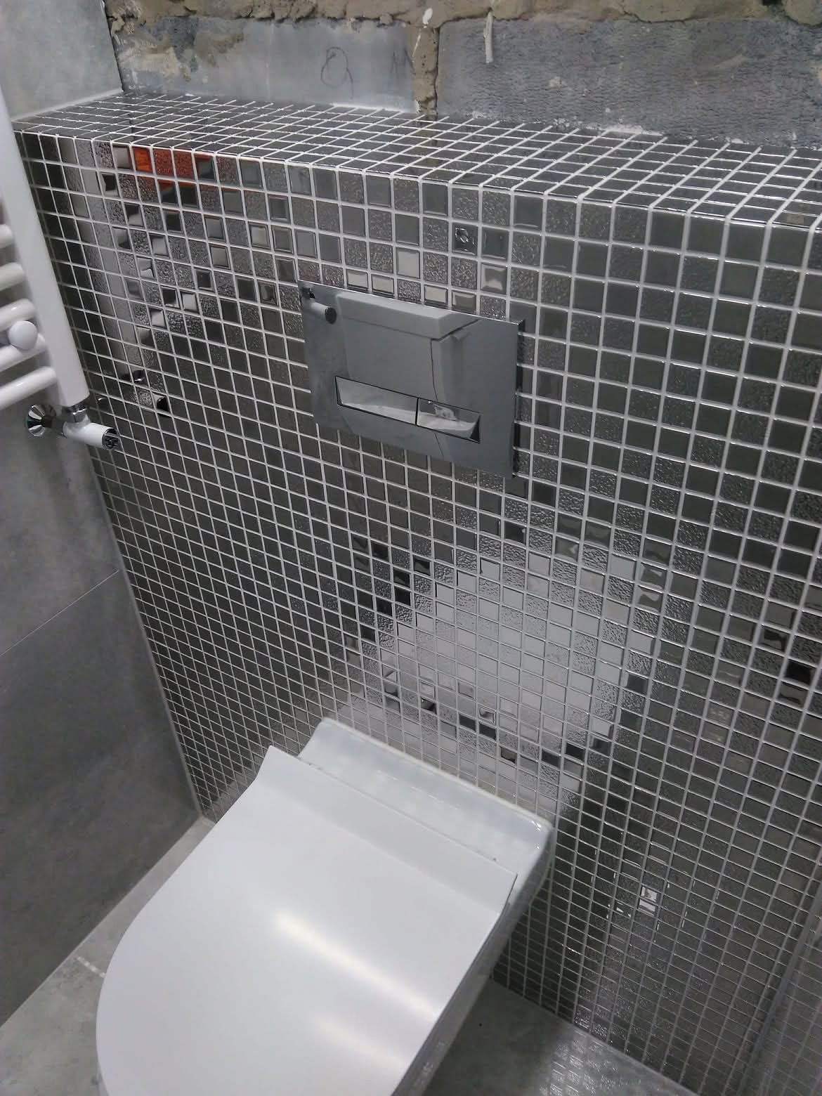 Nowoczesna toaleta z mozaiką w odcieniach szarości i srebrny przycisk spłuczki. Widoczny fragment grzejnika i surowy beton nad płytkami.