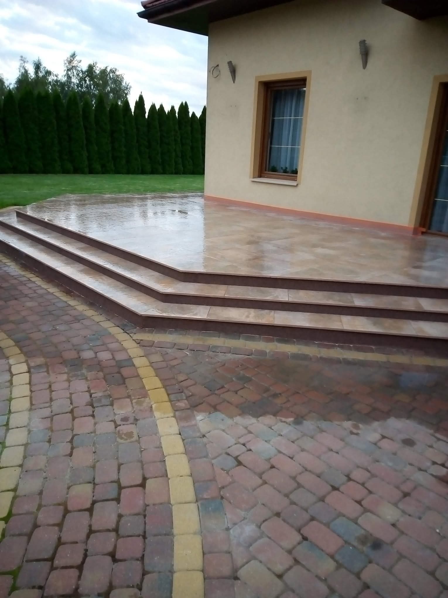 Wykończony taras z trzema stopniami i kostką brukową w odcieniach czerwieni i żółci. Widok na budynek z oknami i zielony żywopłot w tle. Po deszczu, mokra nawierzchnia.
