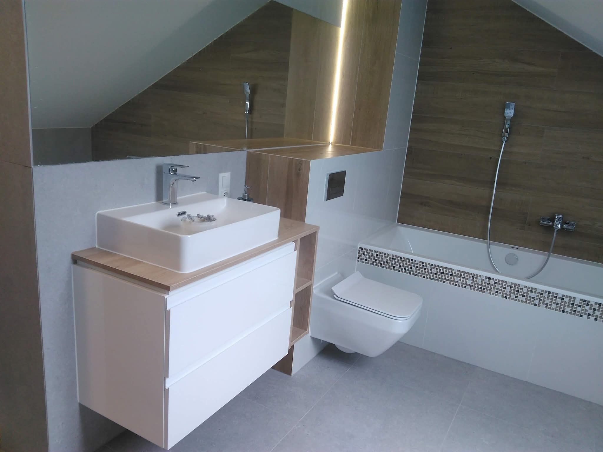 Nowoczesna łazienka na poddaszu z umywalką nablatową, białymi szafkami, toaletą podwieszaną i wanną z mozaiką. Uwagę zwraca oświetlenie LED za lustrem.