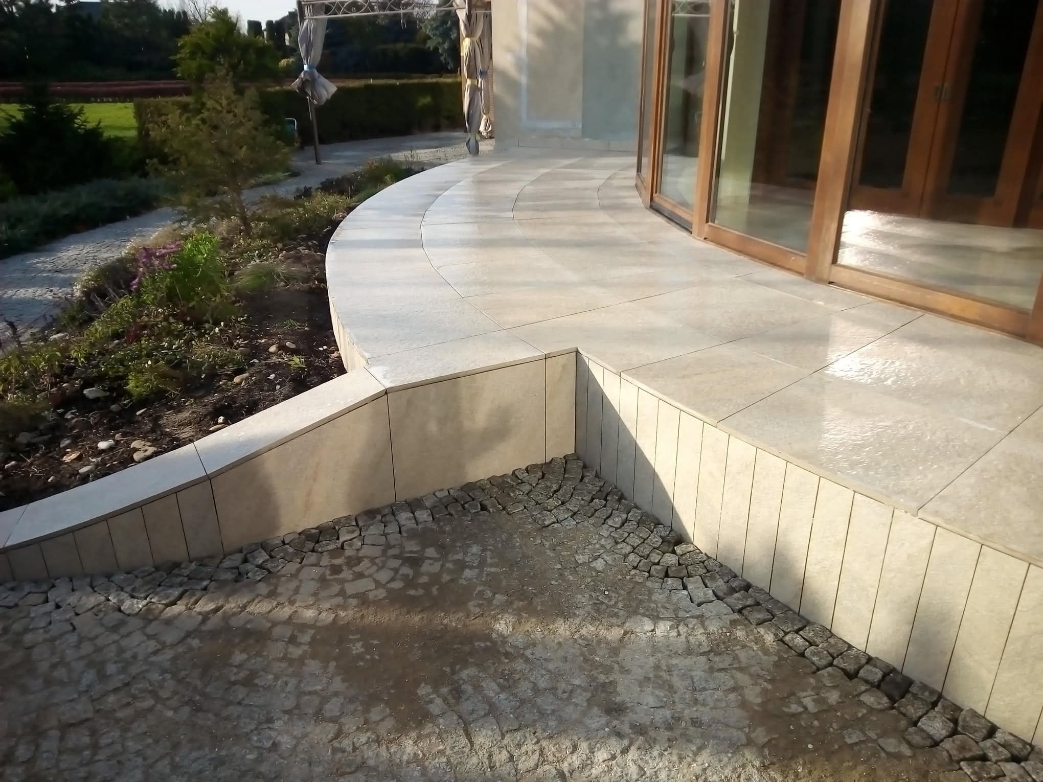 Wykończony taras z jasnymi płytkami i kamienną mozaiką u podstawy, widok na ogród i przeszklone drzwi balkonowe. Estetyczne połączenie materiałów.