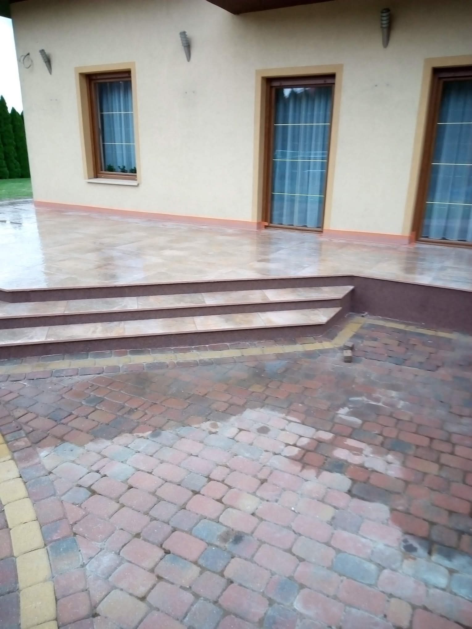 Wykończony taras z płytkami i schodami, obok kostka brukowa. Widoczne okna budynku i oświetlenie zewnętrzne. Całość utrzymana w stonowanej kolorystyce.