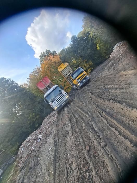 Dwa samochody ciężarowe na nierównym terenie, prawdopodobnie podczas prac ziemnych. Jeden biały z czerwoną skrzynią, drugi żółty. W tle drzewa i błękitne niebo.