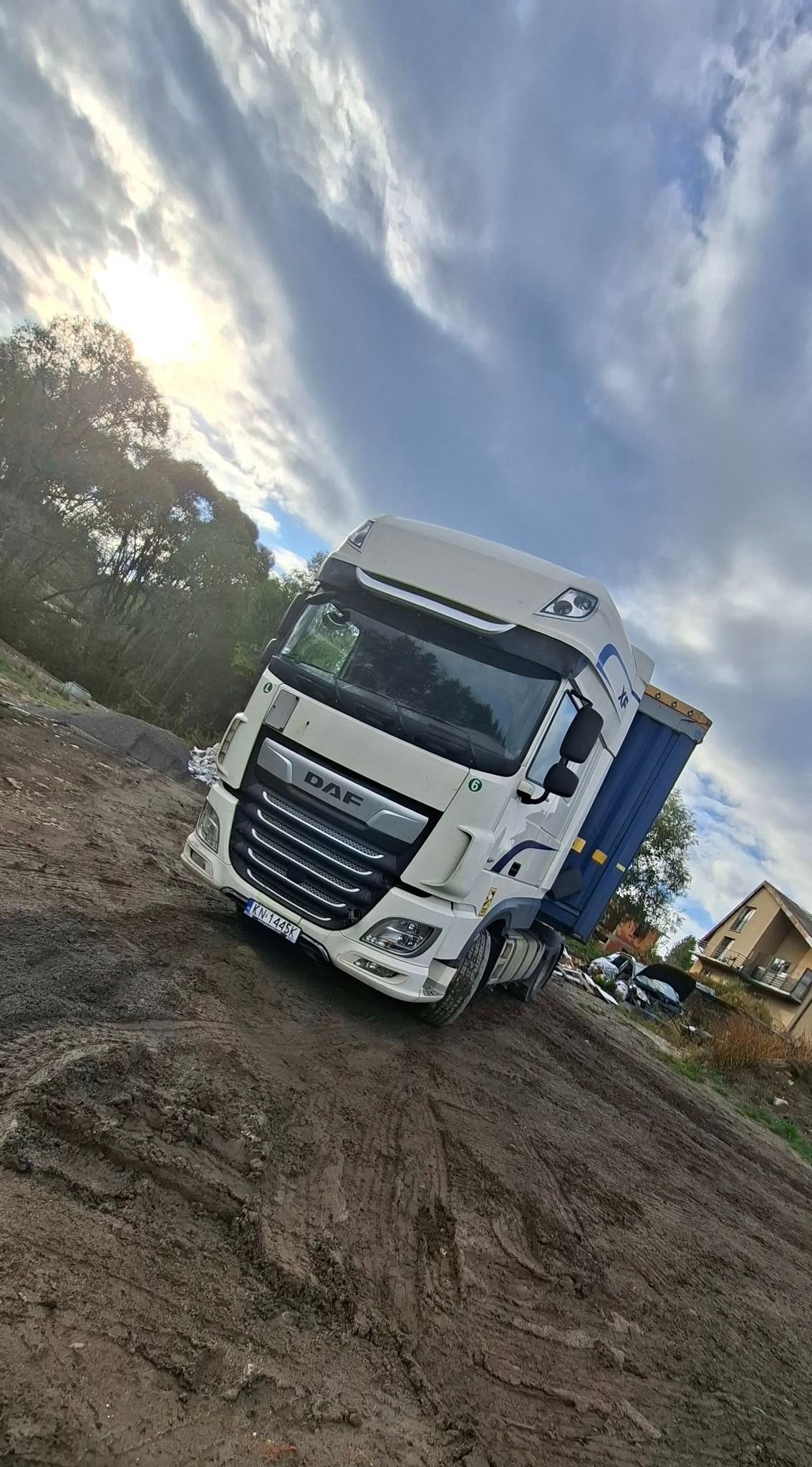 Biała ciężarówka DAF z niebieską skrzynią ładunkową na błotnistym terenie budowy, pod pochmurnym niebem. Widoczny fragment domu w tle.