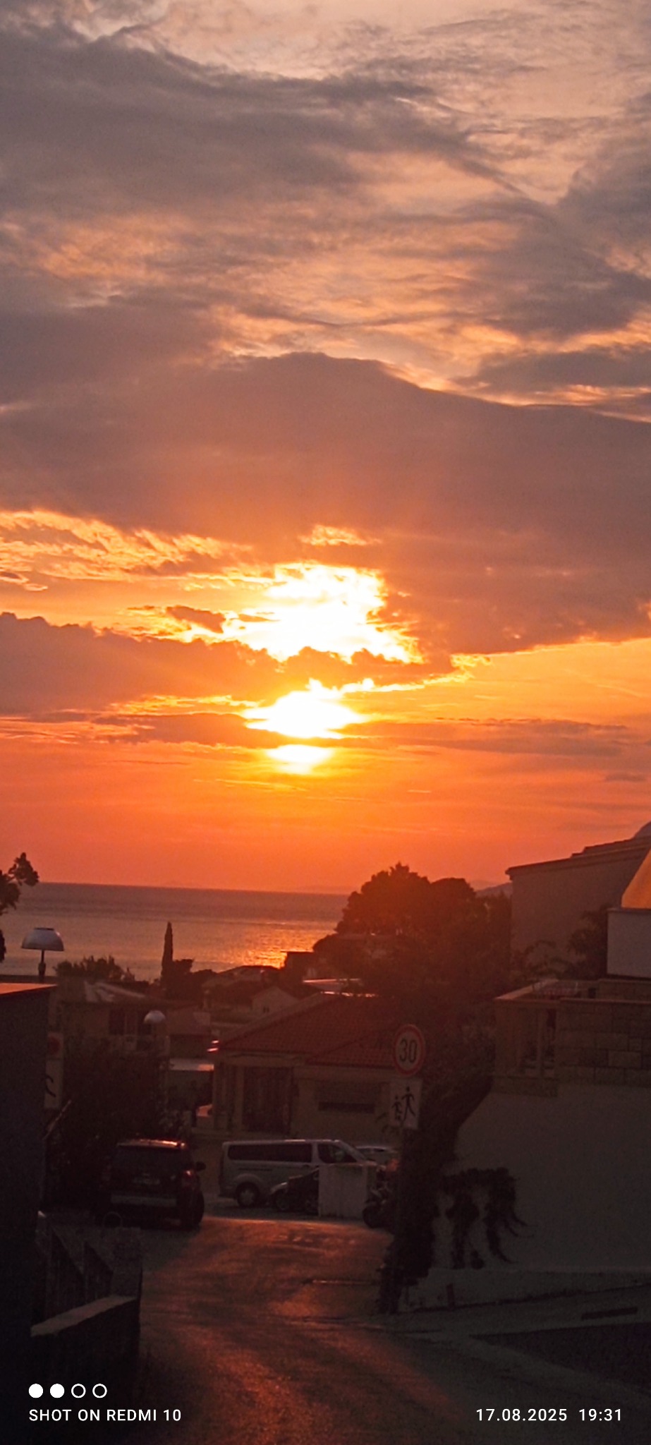 Pomarańczowy zachód słońca nad morzem z widokiem na miasto i ulicę z samochodami. Ujęcie z dołu, z widocznym znakiem drogowym.