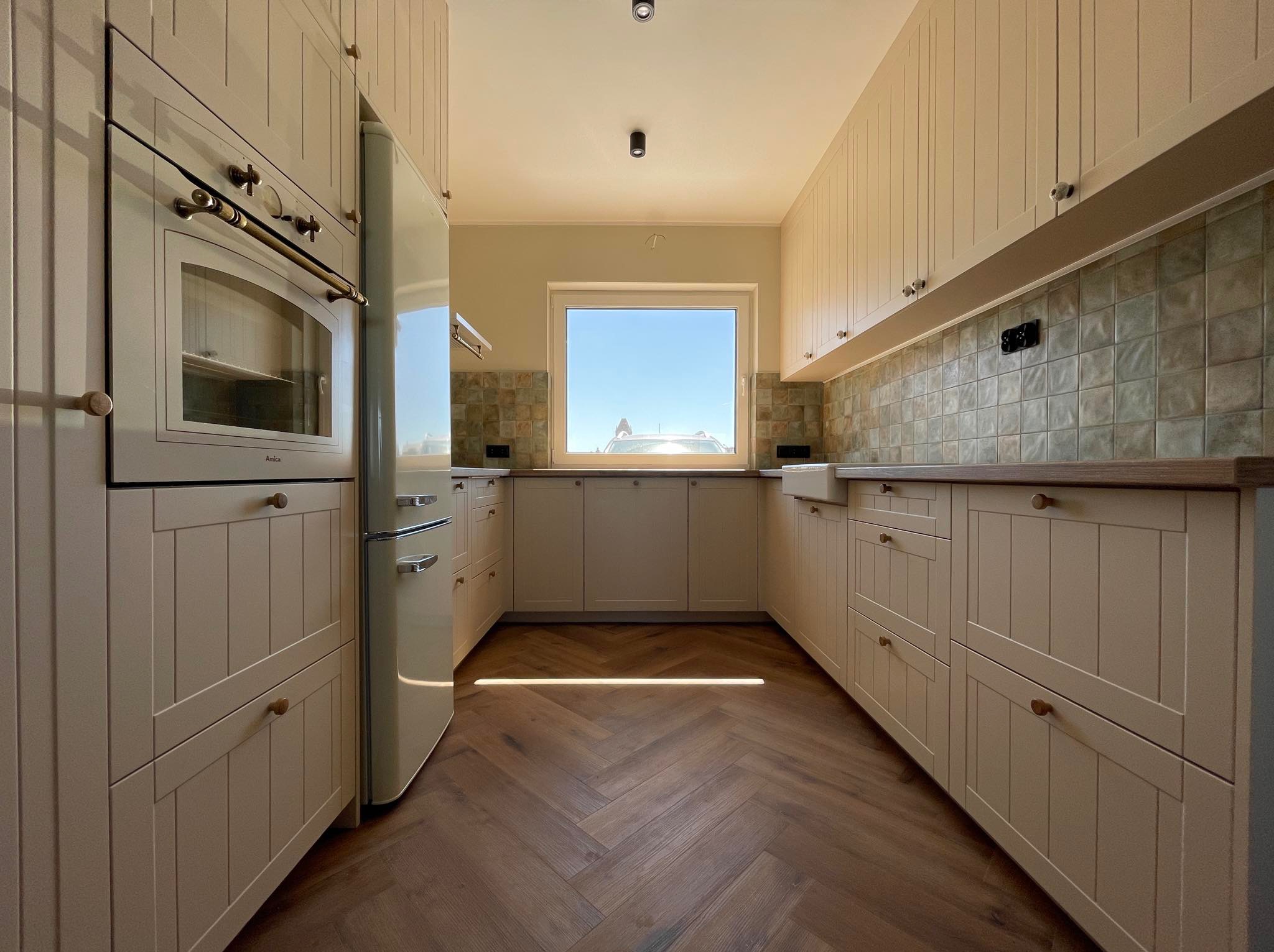 Beżowa kuchnia w stylu retro z drewnianą podłogą w jodełkę. Widoczny piekarnik Amica i lodówka w stylu vintage. Ujęcie z perspektywy wnętrza, z oknem w tle.