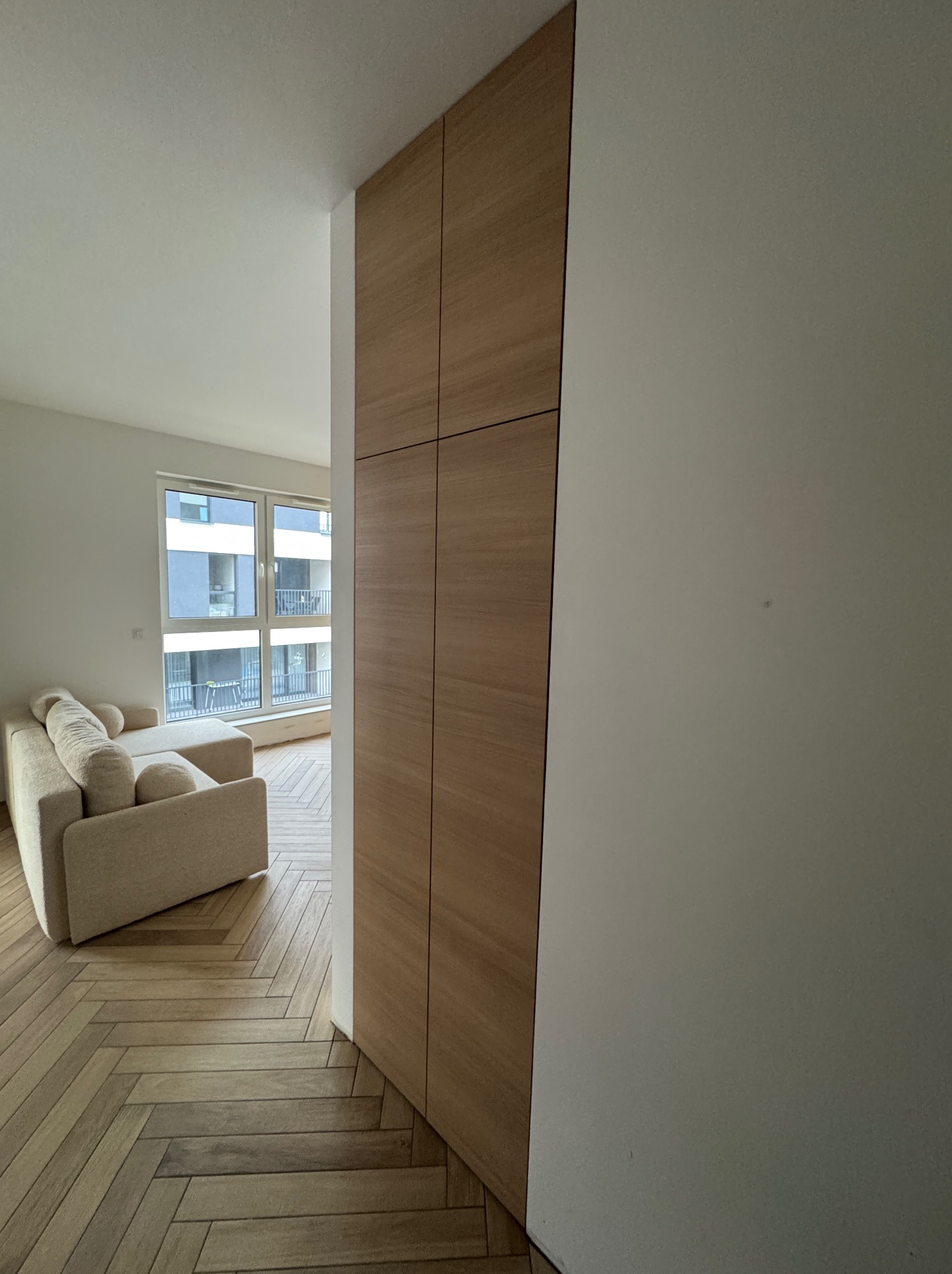 Nowoczesna szafa wnękowa z drewnianymi frontami, wkomponowana w jasne wnętrze mieszkania z parkietem w jodełkę i sofą. Minimalistyczny design, naturalne materiały.