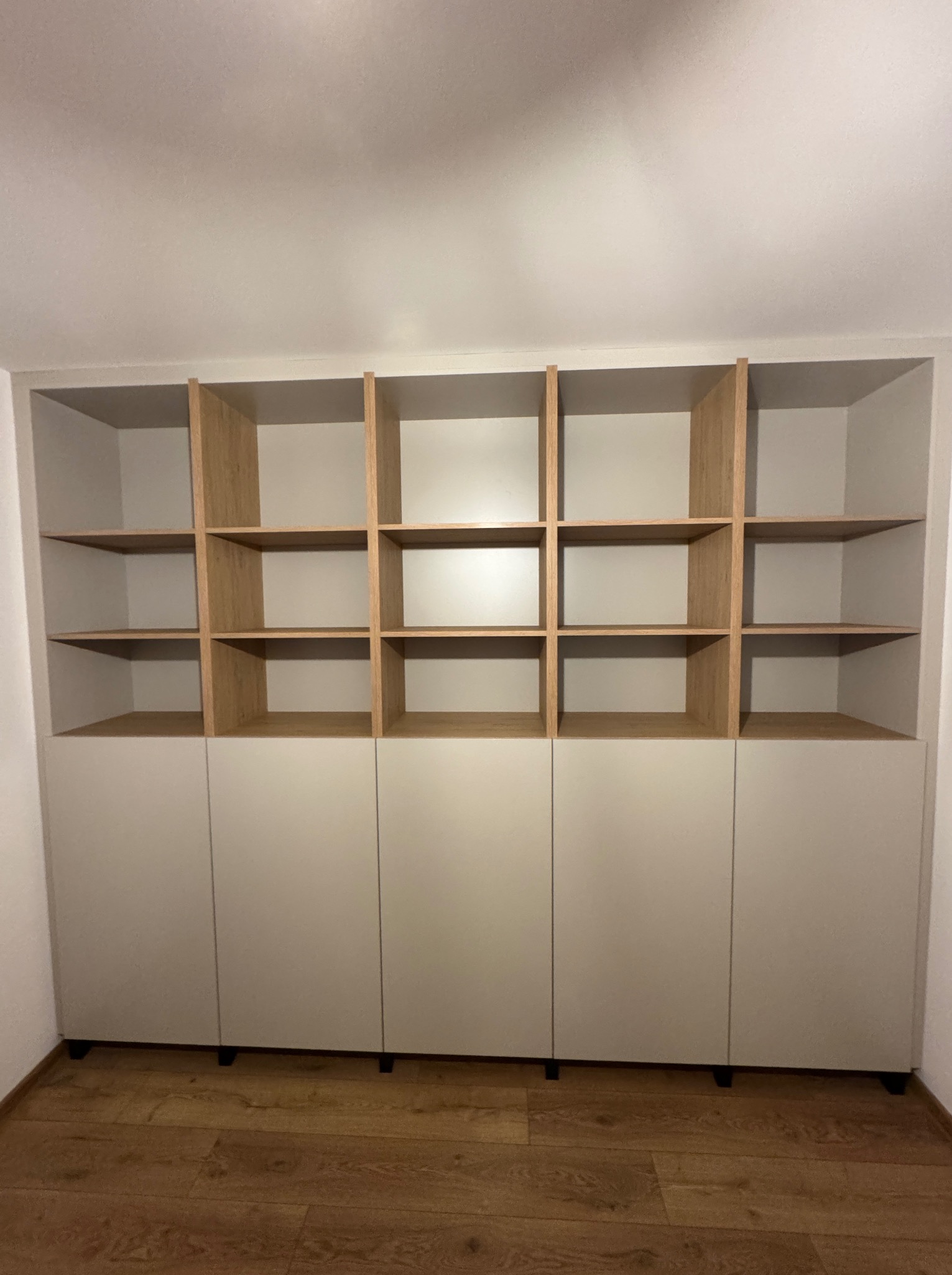 Biało-drewniana szafa wnękowa z otwartymi półkami i zamkniętymi szafkami na dole, na drewnianej podłodze. Minimalistyczny design.