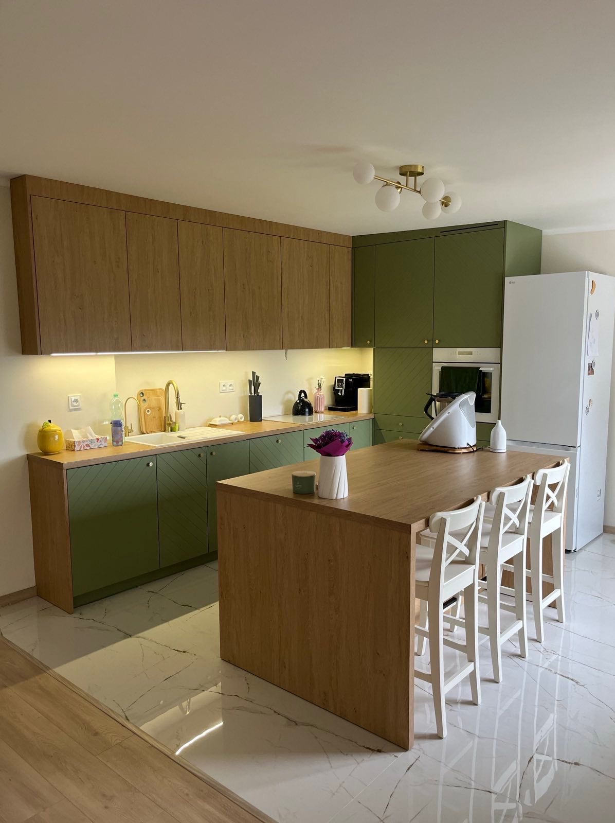 Nowoczesna kuchnia z wyspą: fronty w odcieniach zieleni i drewna, białe hokery, marmurowe podłogi. Oświetlenie LED pod szafkami. Minimalistyczny design.