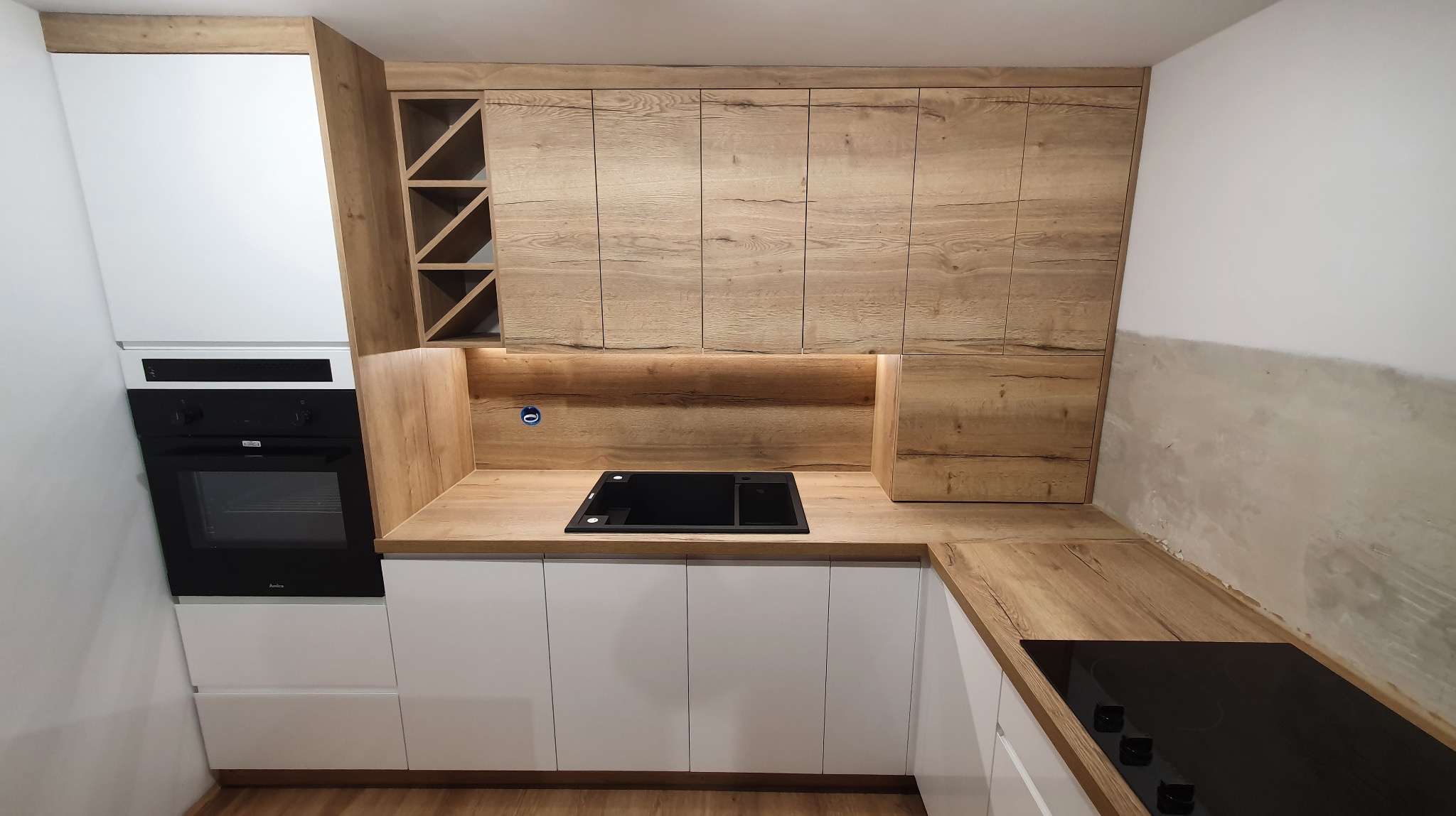Nowoczesna kuchnia z białymi szafkami dolnymi i drewnopodobnymi górnymi, z czarnym piekarnikiem i płytą. Widoczny regał na wino i oświetlenie podszafkowe.