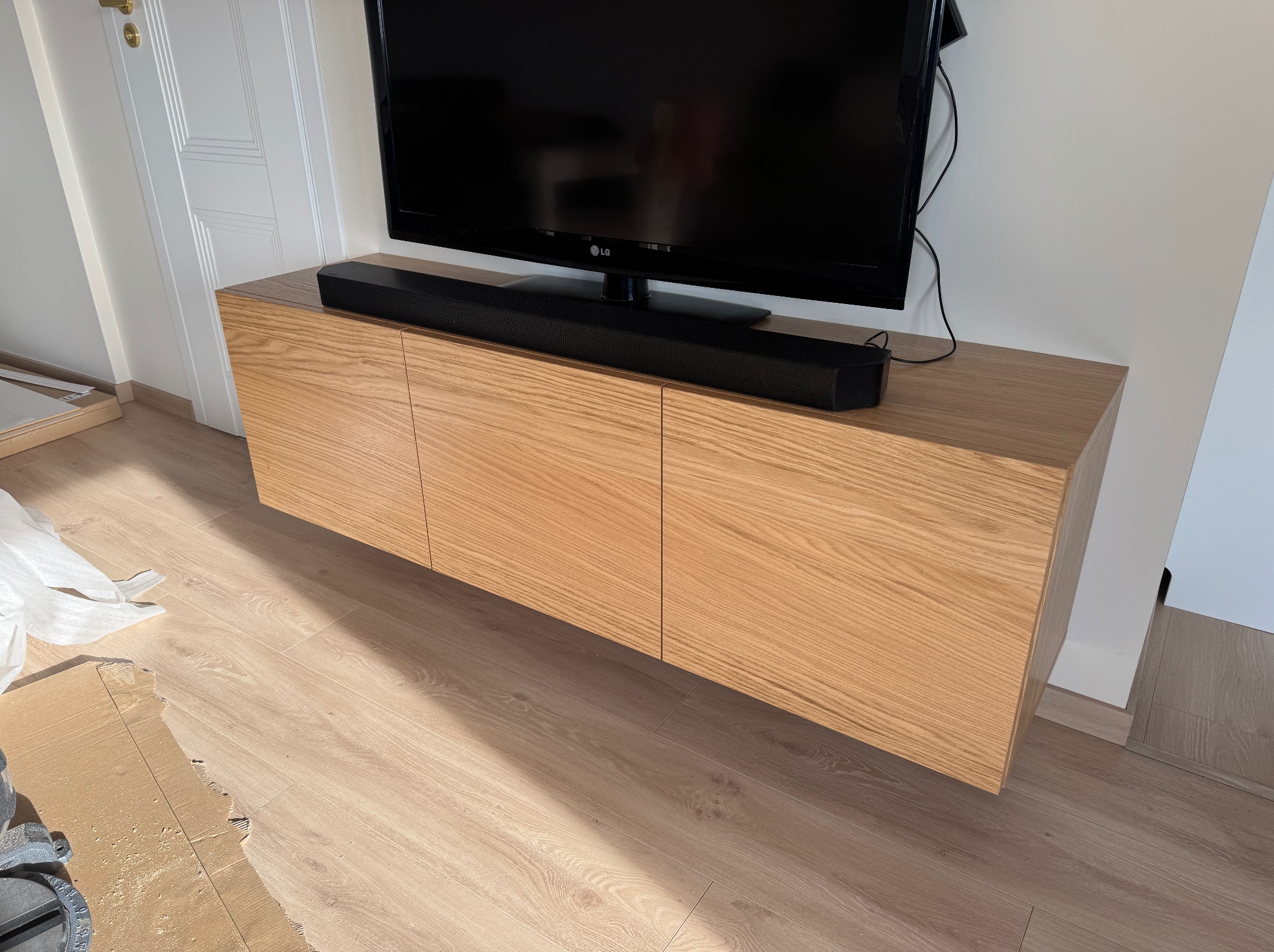 Drewniana szafka RTV z frontami bez uchwytów, na której stoi telewizor i soundbar. Jasne drewno, minimalistyczny design, nowoczesne wnętrze.