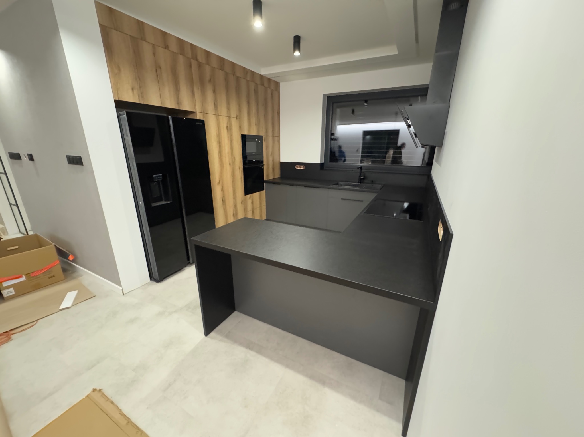 Nowoczesna kuchnia w odcieniach szarości i drewna z czarnym sprzętem AGD. Minimalistyczny design, oświetlenie punktowe i duża lodówka side-by-side.