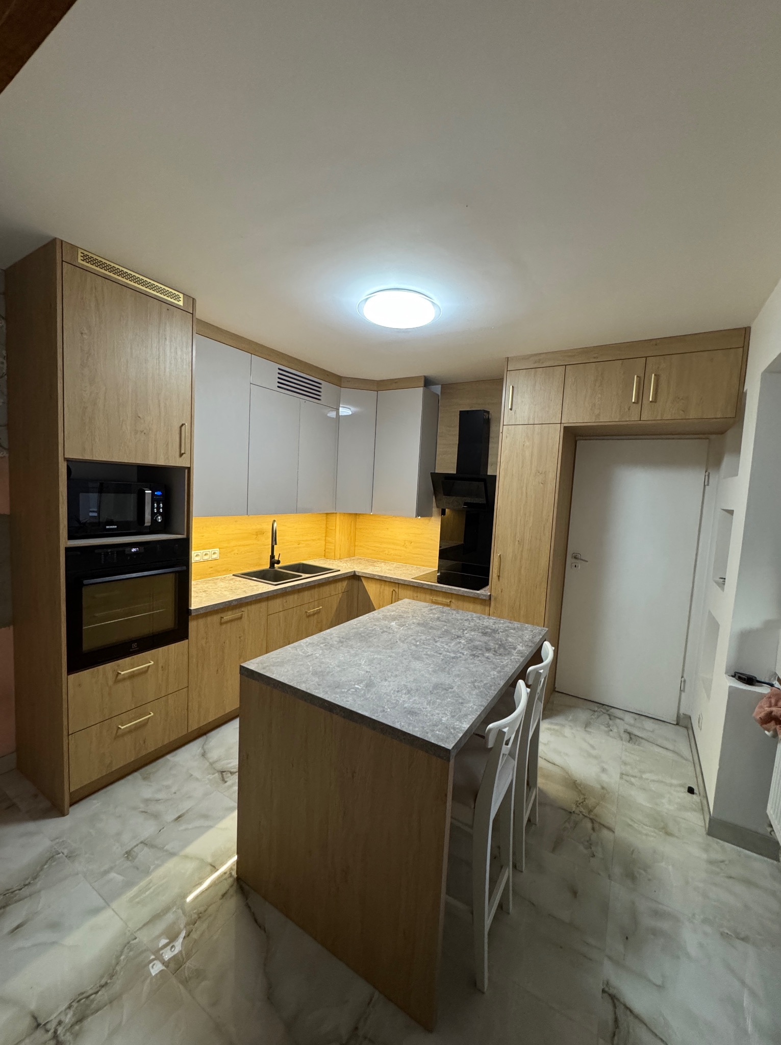 Jasna kuchnia z wyspą i marmurową podłogą. Fronty szafek w odcieniach drewna i szarości. Oświetlenie LED pod szafkami górnymi. Nowoczesny design.