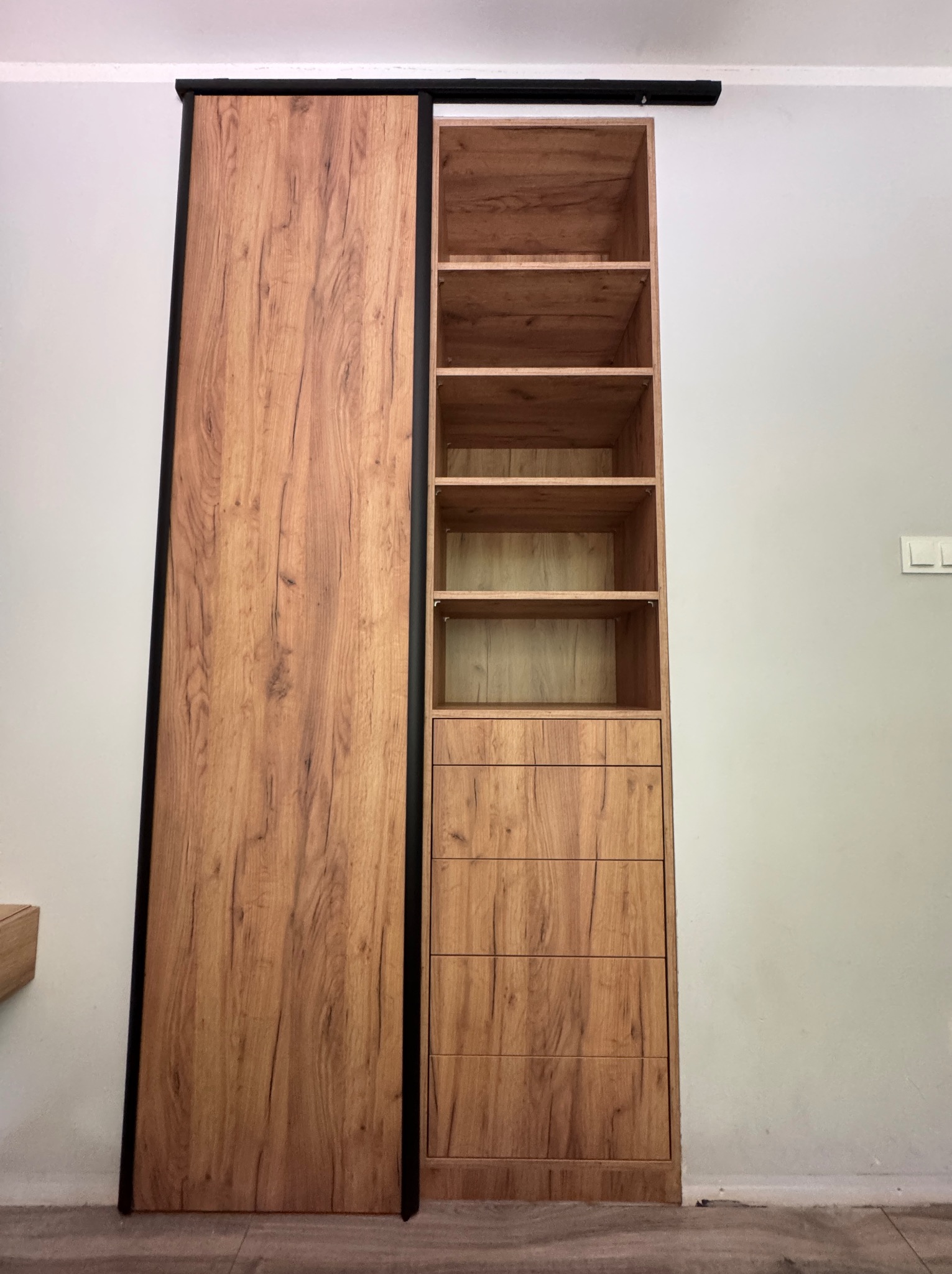 Drewniana szafa wnękowa z przesuwnymi drzwiami i otwartymi półkami, wykończona w ciepłym odcieniu drewna z czarnymi detalami, na tle jasnej ściany.