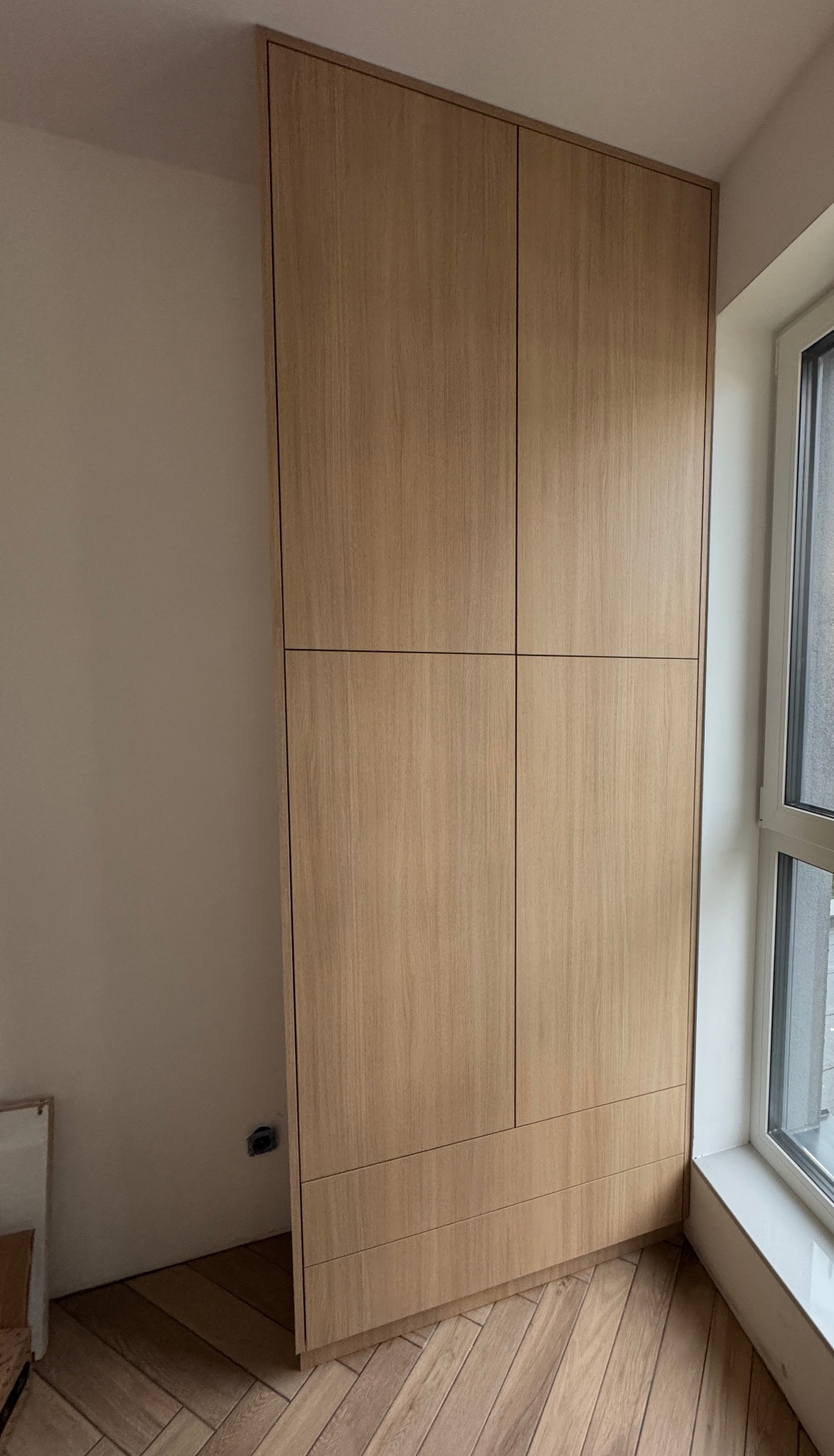 Jasna, drewniana szafa wnękowa z czterema drzwiami i trzema szufladami, wkomponowana w narożnik pokoju z parkietem ułożonym w jodełkę, obok okna.