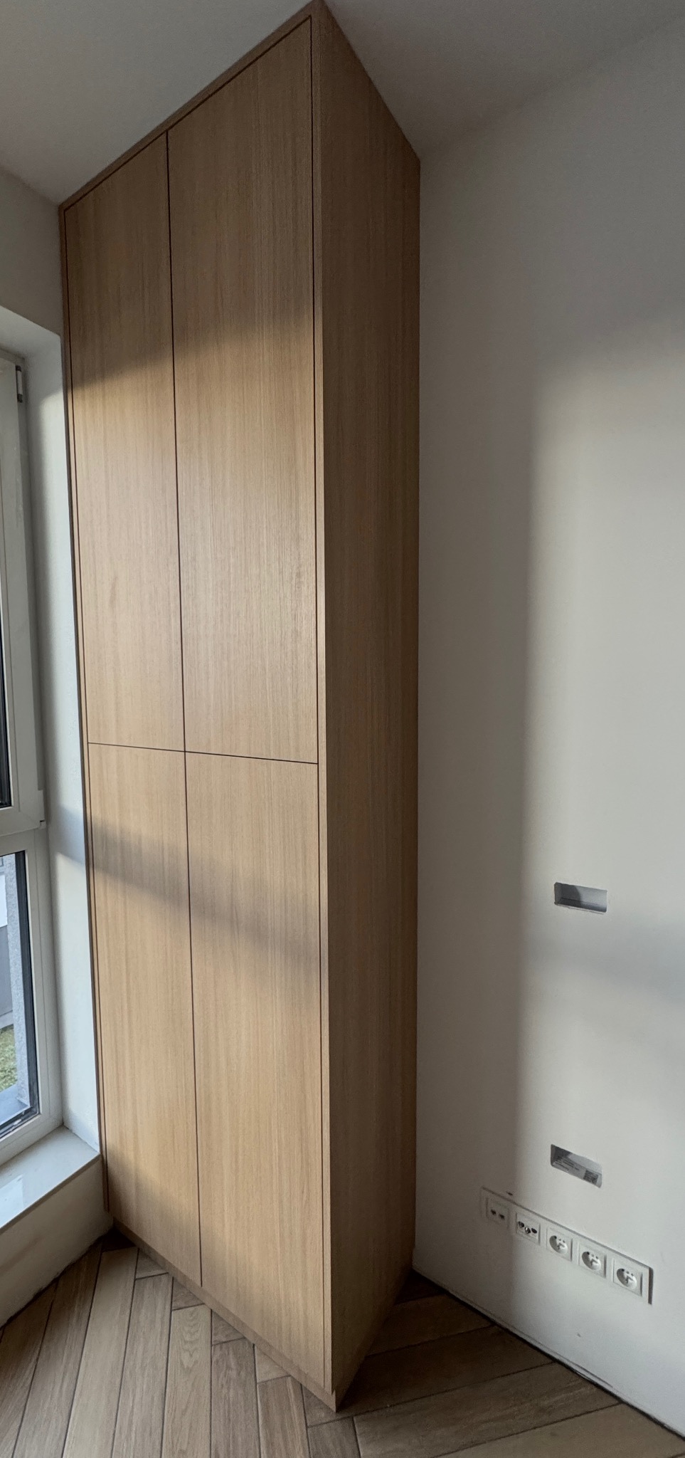 Wysoka, drewniana szafa wnękowa z czterema drzwiami, z delikatnym usłojeniem, dopasowana do jasnego wnętrza z drewnianą podłogą i oknem po lewej stronie.