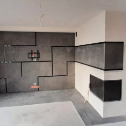 Artur Jałowiecki - Pokój w trakcie wykańczania: ściana z płyt w geometryczne wzory, przygotowane miejsce na TV, obudowa kominka. Widoczne narzędzia i materiały budowlane.