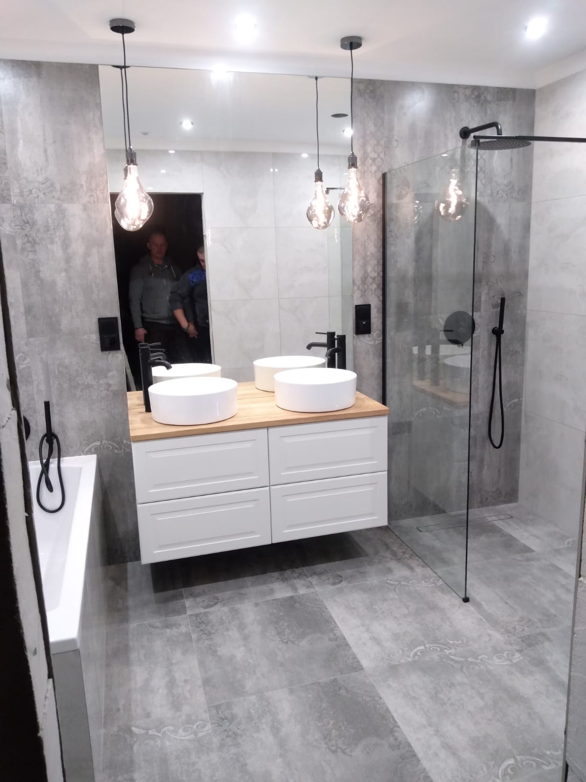 Nowoczesna łazienka w szarościach: biała szafka z umywalkami nablatowymi, kabina prysznicowa ze szklaną ścianą, dekoracyjne płytki podłogowe i wiszące lampy.
