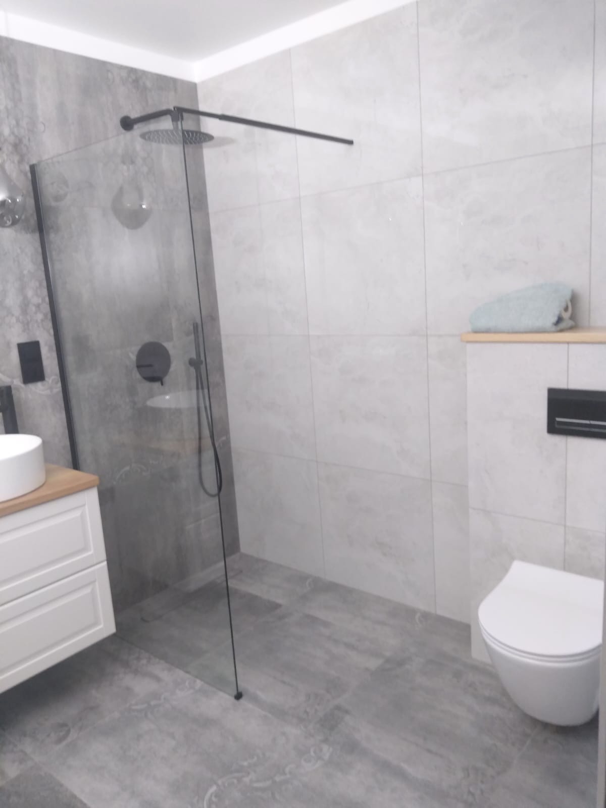 Nowoczesna łazienka z kabiną typu walk-in, szare płytki imitujące kamień, biała umywalka nablatowa i toaleta. Minimalistyczny design z czarnymi akcentami.