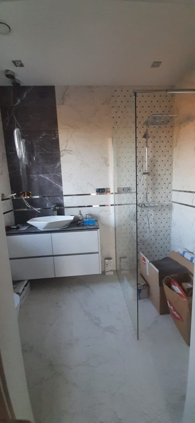 Nowoczesna łazienka w trakcie wykańczania: biała umywalka na szafce, szklana kabina prysznicowa, marmurowe płytki na ścianach z dekoracyjnym wzorem.