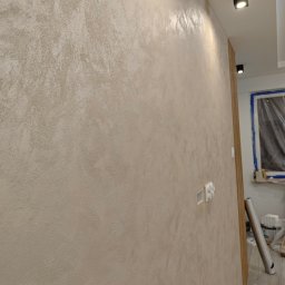 BUD-RAF Ślebocki Rafał - Ściana z dekoracyjną, beżową strukturą, oświetlona punktowo. Widoczne gniazdka elektryczne i fragment okna z folią ochronną. Efektowna faktura na ścianie.