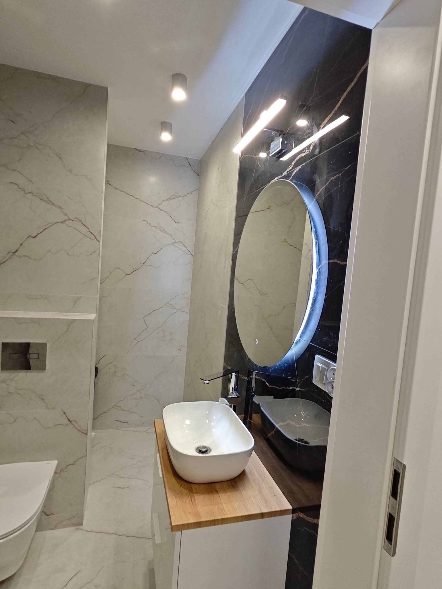 Nowoczesna łazienka z marmurowymi płytkami, owalnym lustrem z podświetleniem LED i umywalką nablatową na drewnianym blacie. Minimalistyczny design, eleganckie wykończenie.