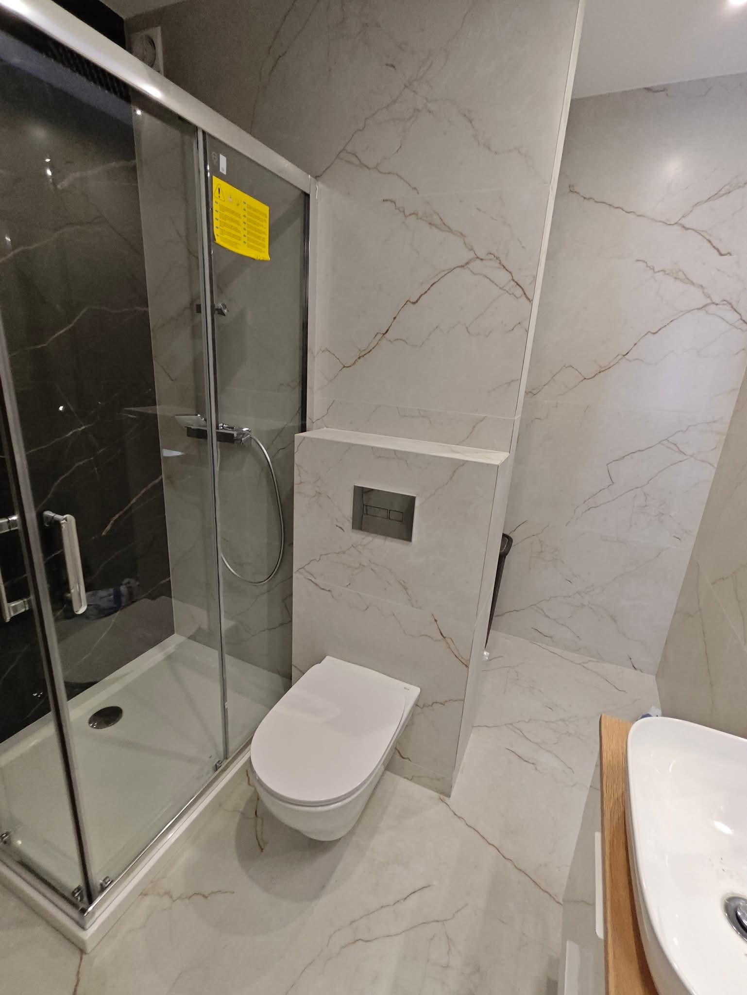 Wykończona łazienka w Kutno: kabina prysznicowa ze szklanymi drzwiami, podwieszana toaleta i umywalka nablatowa na drewnianym blacie, marmurowe płytki na ścianach i podłodze.