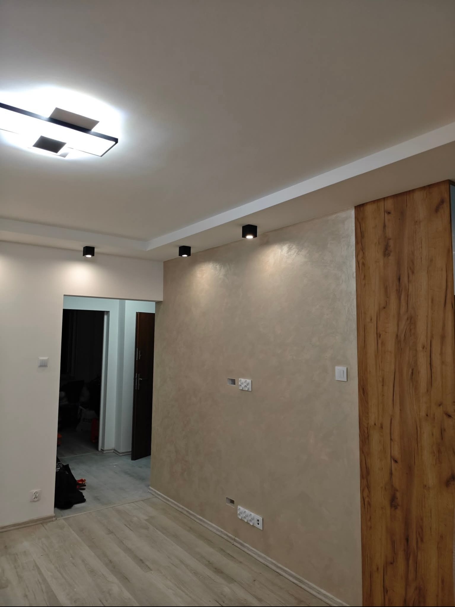 Wykończone wnętrze: ściana z dekoracyjną fakturą w beżach, drewniana okładzina, nowoczesne oświetlenie sufitowe i podłoga z paneli. Widok na otwarte przejście do innego pomieszczenia.