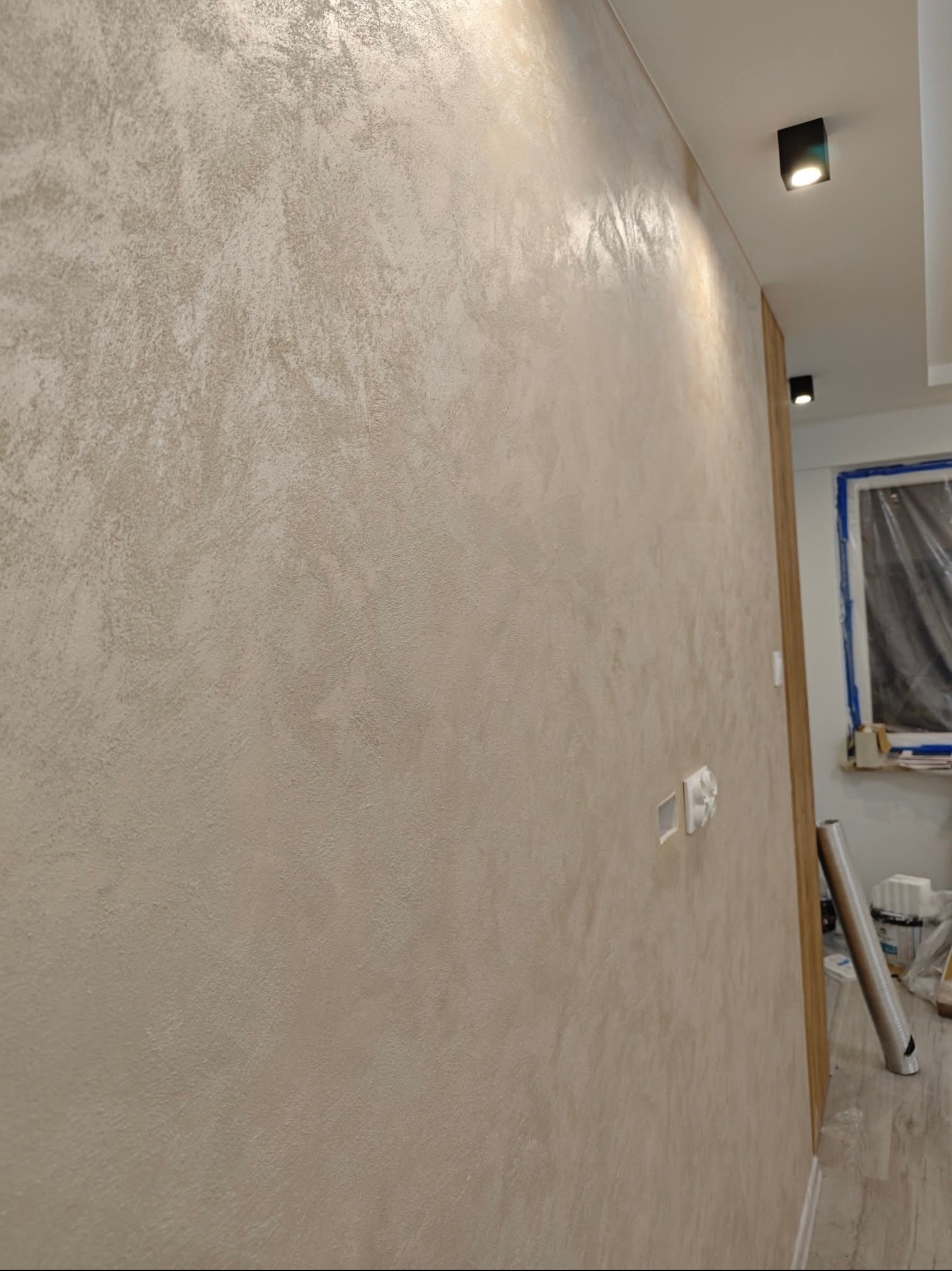 Ściana z dekoracyjną, beżową strukturą, oświetlona punktowo. Widoczne gniazdka elektryczne i fragment okna z folią ochronną. Efektowna faktura na ścianie.