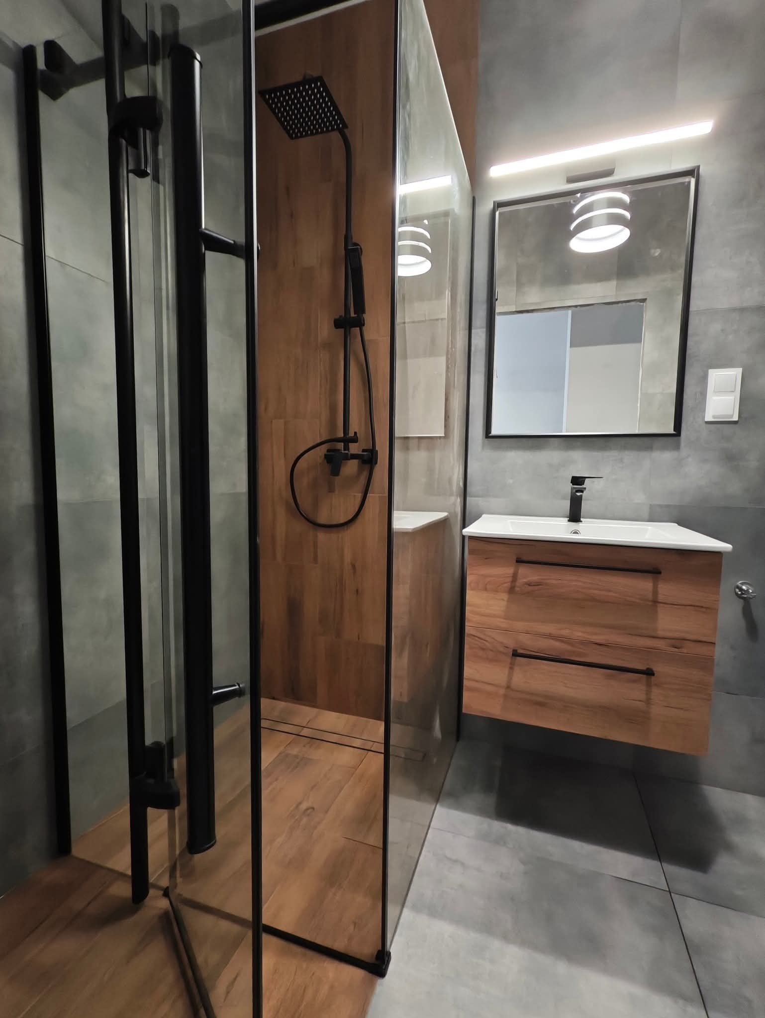 Nowoczesna łazienka z kabiną prysznicową typu walk-in, drewnianą ścianą i szafką pod umywalką z drewnianym frontem. Szare płytki na ścianach i podłodze.