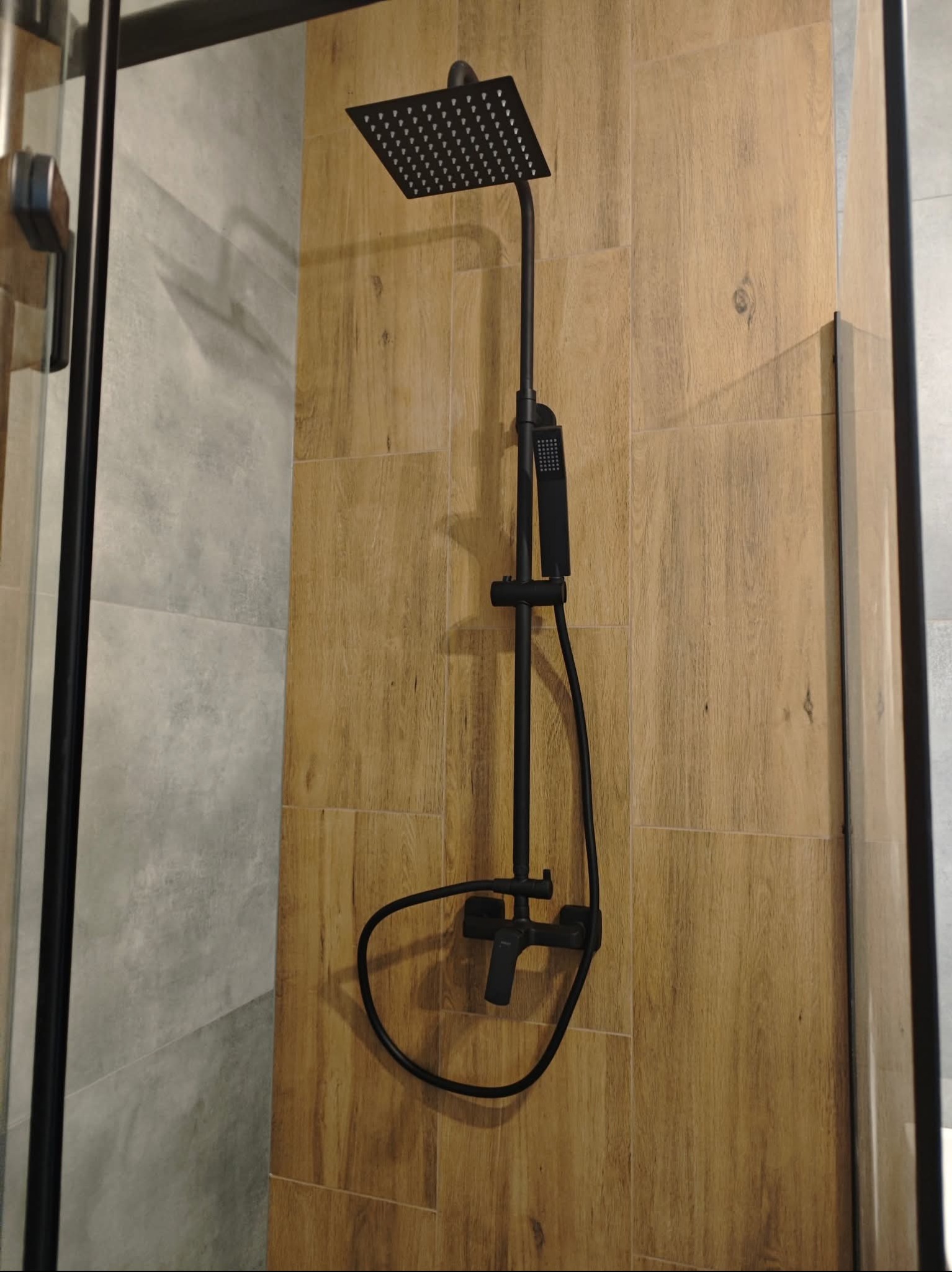 Nowoczesny, czarny zestaw prysznicowy z deszczownicą na ścianie wyłożonej płytkami imitującymi drewno, w kabinie prysznicowej z elementami szklanymi.