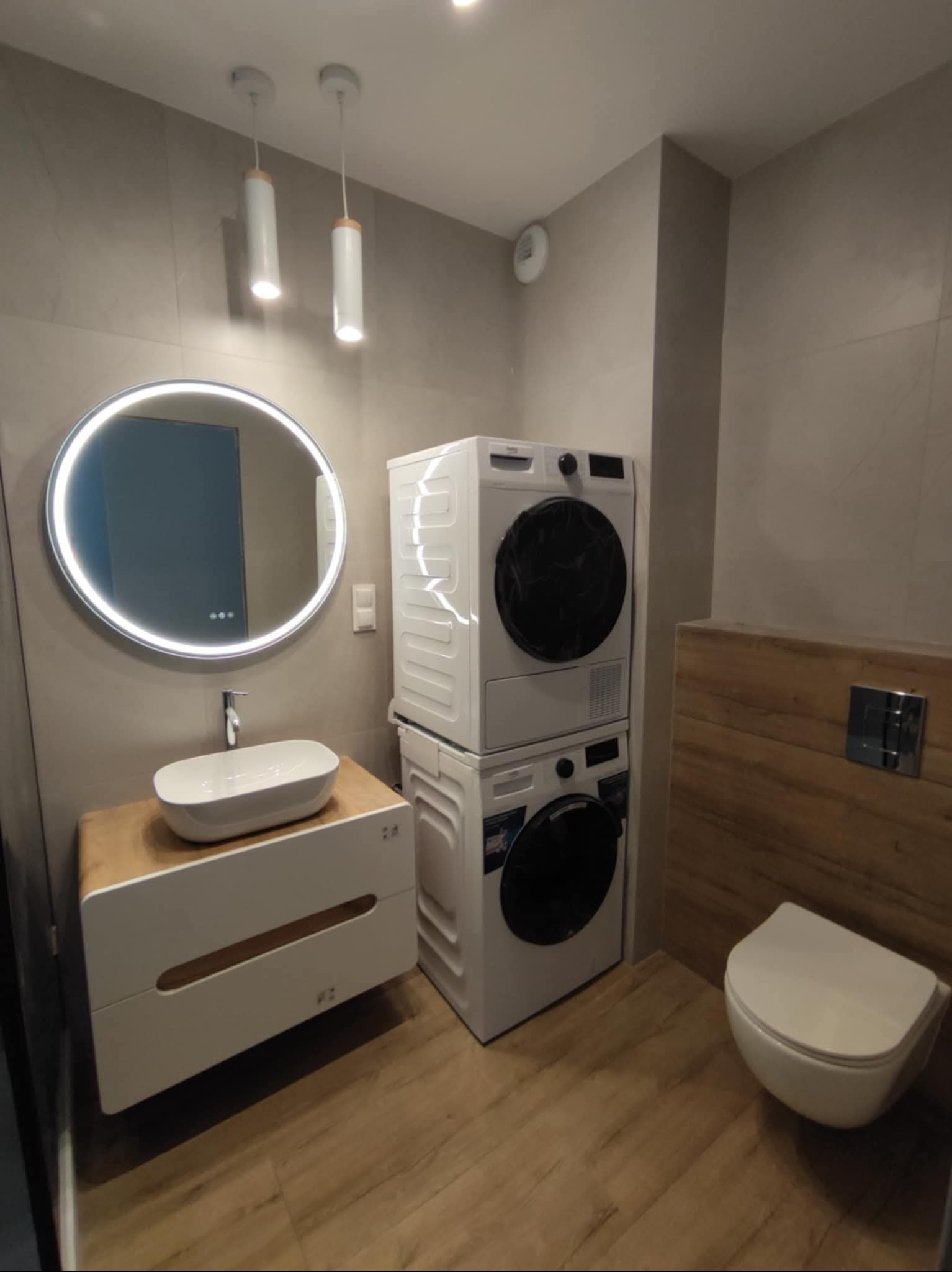 Nowoczesna łazienka z podświetlanym lustrem, umywalką na szafce z drewnianym blatem, toaletą i pralko-suszarką w zabudowie. Minimalistyczny design, neutralna kolorystyka.