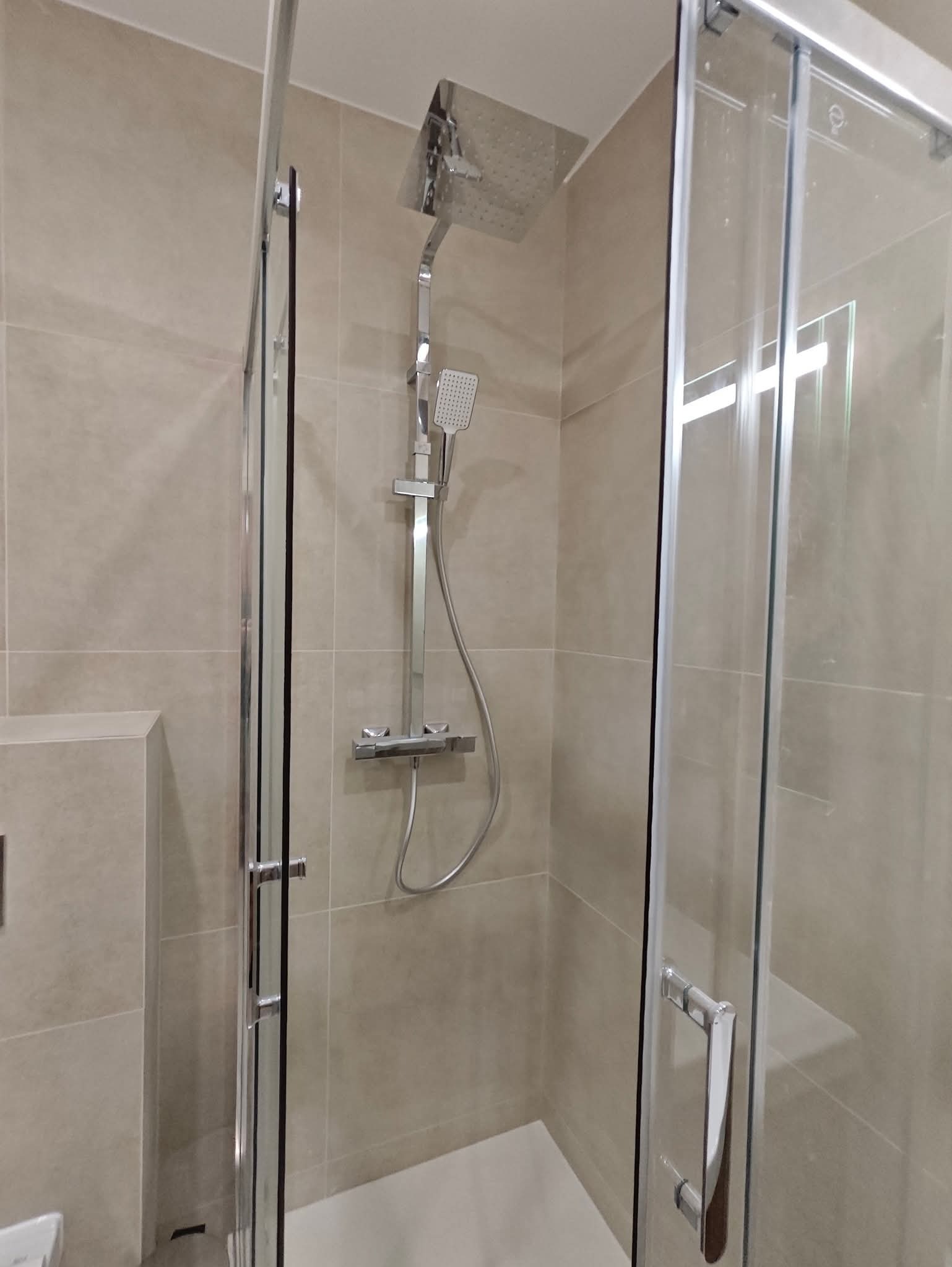 Nowoczesna kabina prysznicowa z deszczownicą i szklanymi drzwiami przesuwnymi, wykończona beżowymi płytkami. Minimalistyczny design, chromowane elementy.