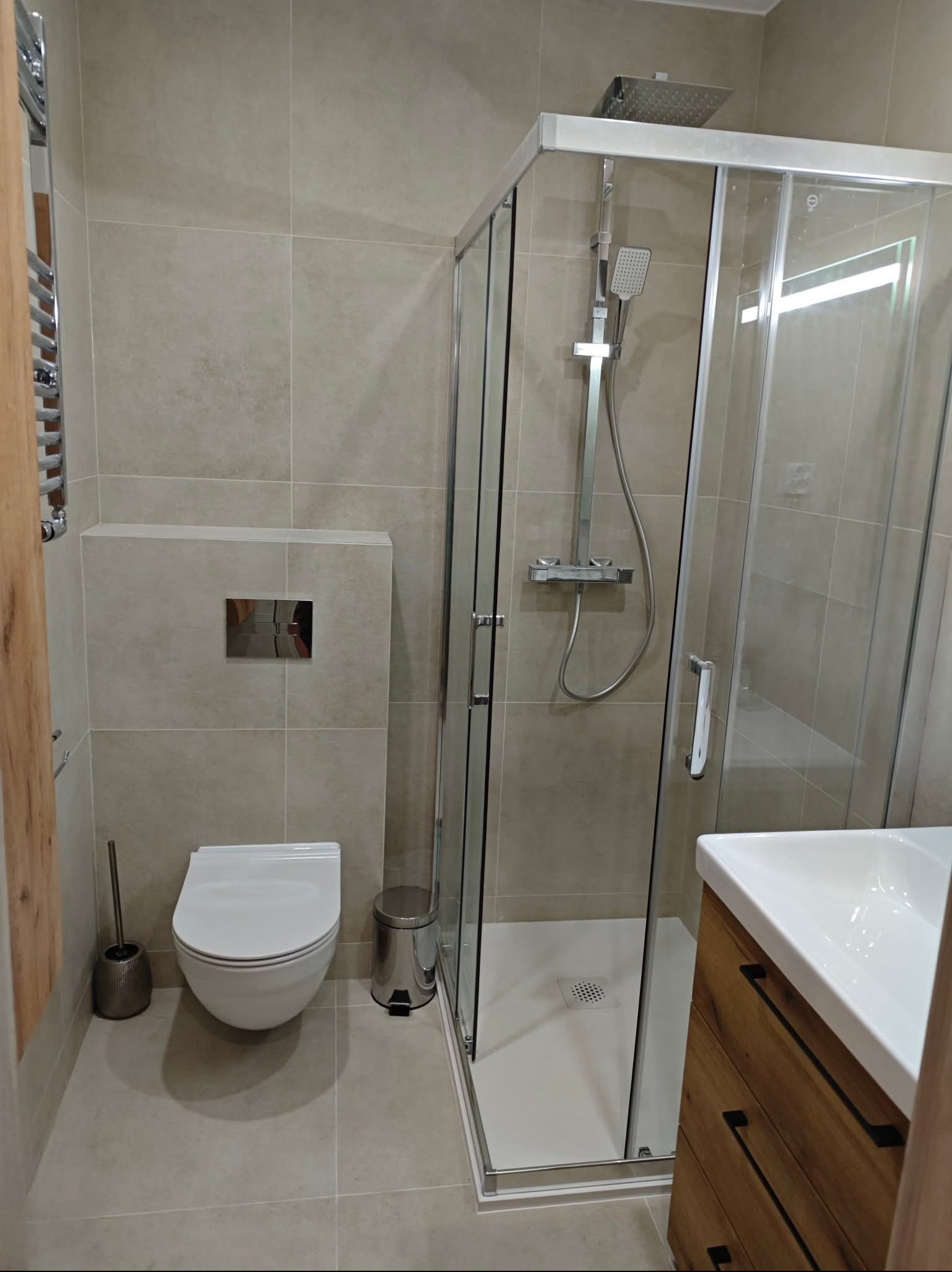 Nowoczesna łazienka z kabiną prysznicową typu walk-in, podwieszaną toaletą i drewnianą szafką pod umywalką. Beżowe płytki na ścianach i podłodze. Minimalistyczny design.