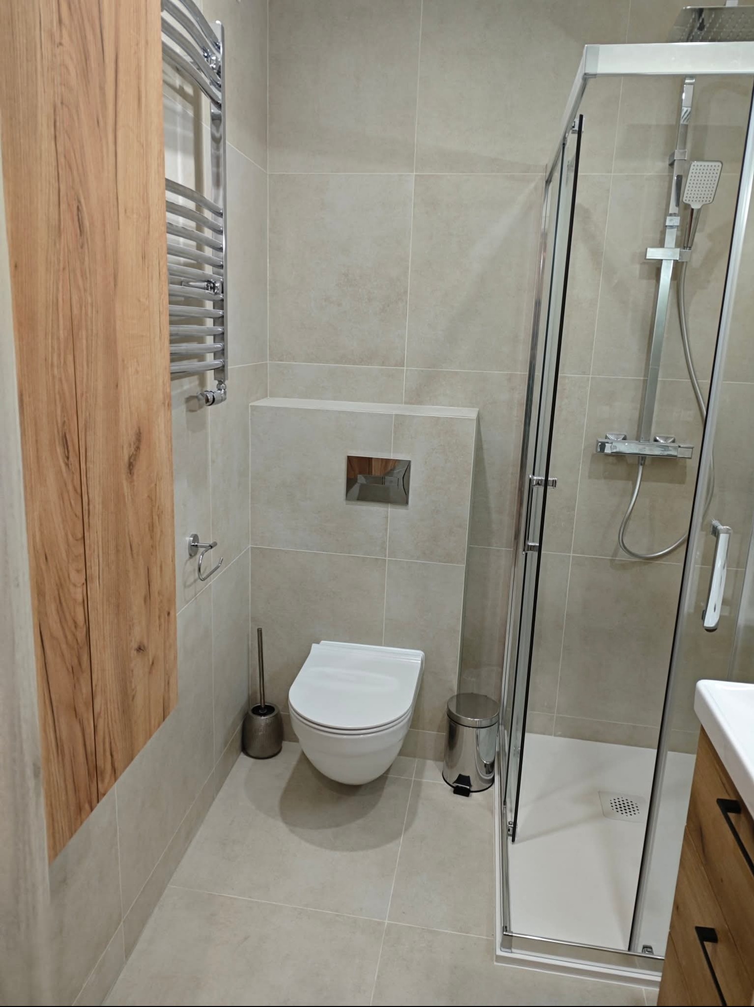 Nowoczesna łazienka z kabiną prysznicową, toaletą podwieszaną i grzejnikiem drabinkowym. Jasne płytki, drewniane akcenty i chromowane dodatki tworzą spójną całość.
