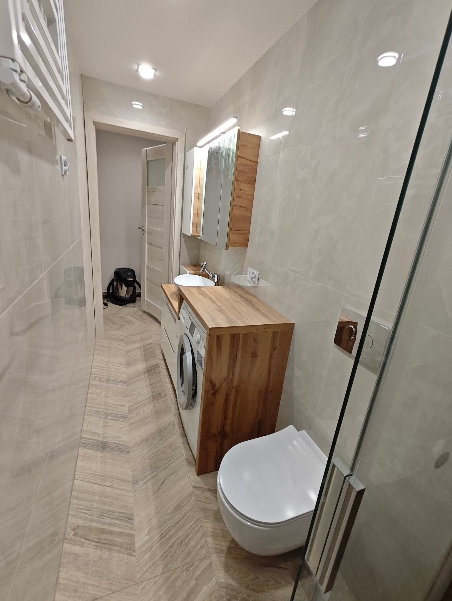 Jasna łazienka po remoncie: drewniany blat z umywalką nad pralką, toaleta, kabina prysznicowa, płytki imitujące drewno na podłodze, drzwi z przeszkleniem.