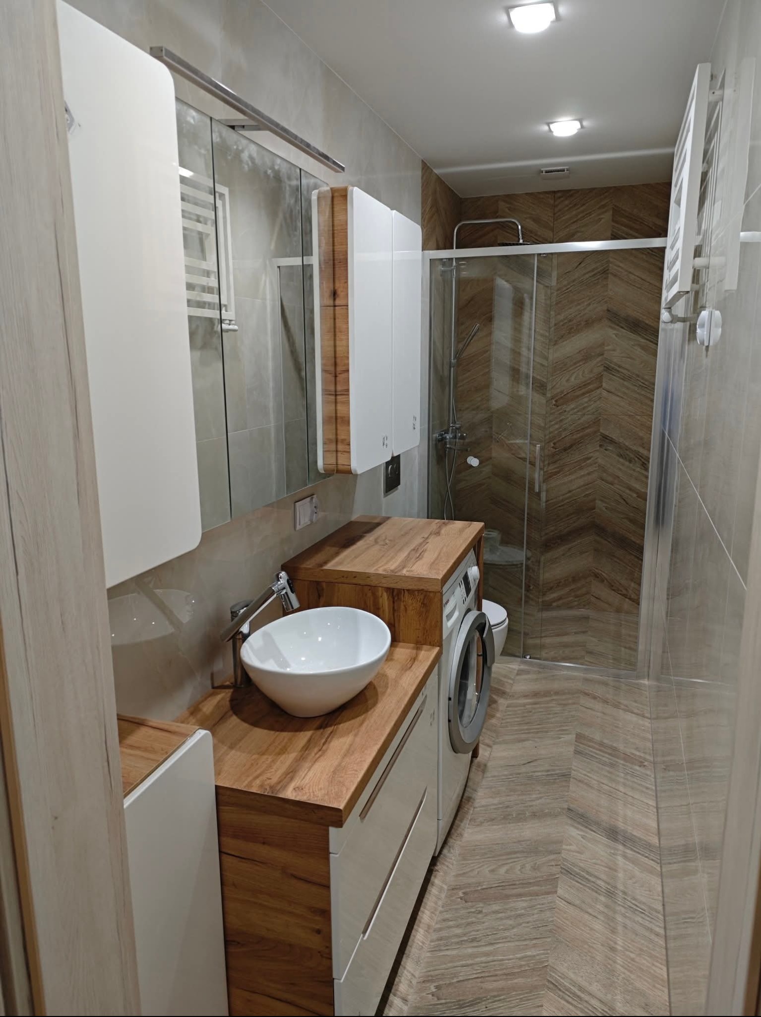 Nowoczesna łazienka z drewnianymi akcentami: blat z umywalką, szafki, pralka i kabina prysznicowa z brązowymi płytkami ułożonymi w jodełkę. Całość w jasnych, stonowanych barwach.