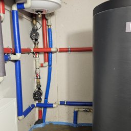 Tetiana Senkiv - Złożona instalacja grzewcza z rurami w izolacji (czerwone i niebieskie) oraz zbiornikiem akumulacyjnym. Widoczne pompy obiegowe i fragment okna w tle.