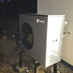 Tetiana Senkiv - Szara pompa ciepła Kensol HVAC zamontowana na zewnątrz budynku, widoczne rury przyłączeniowe i betonowe podstawy. W tle las i wieczorne niebo.